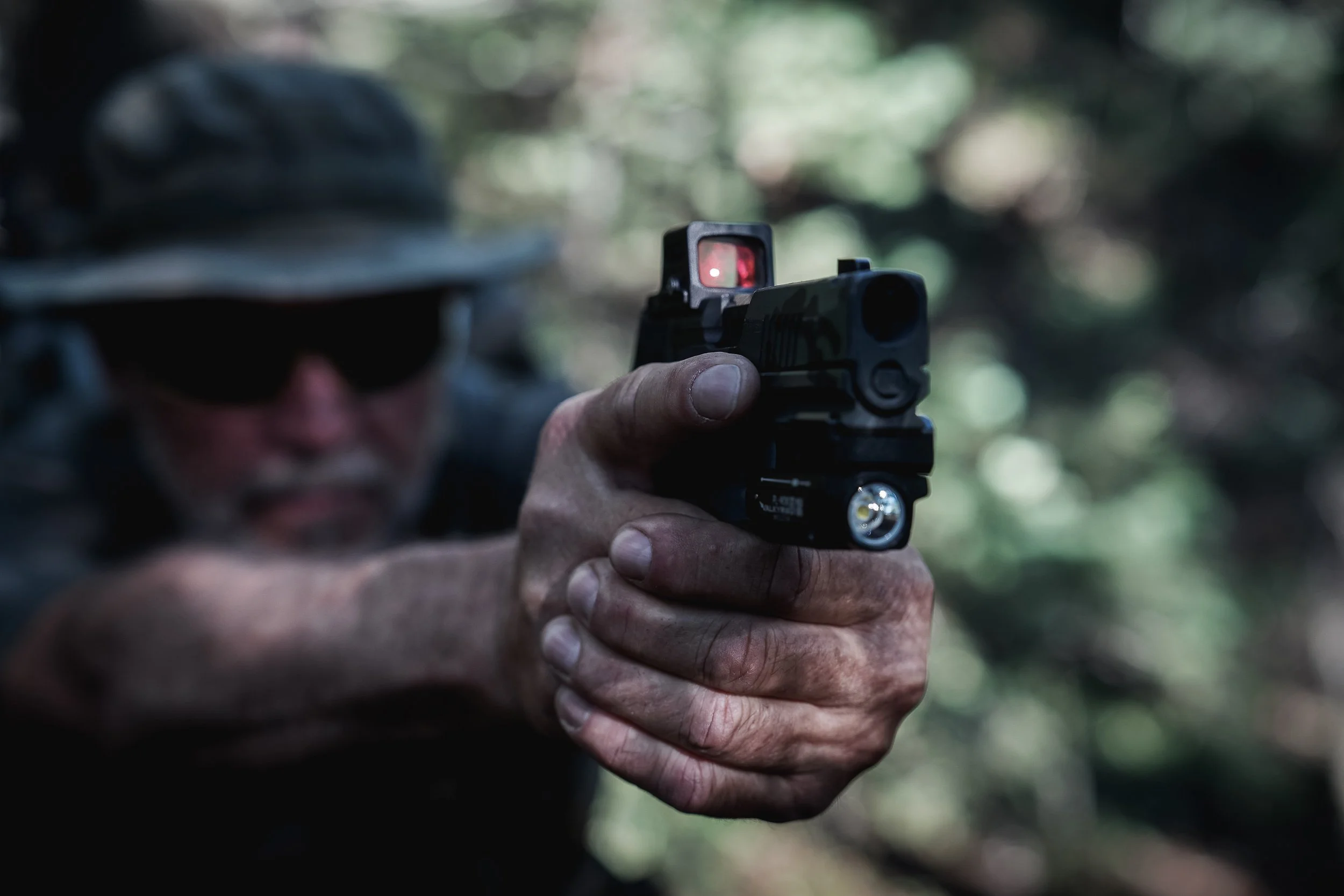 A person wearing a hat and dark sunglasses aiming a handgun outdoors, with a blurred forest background.