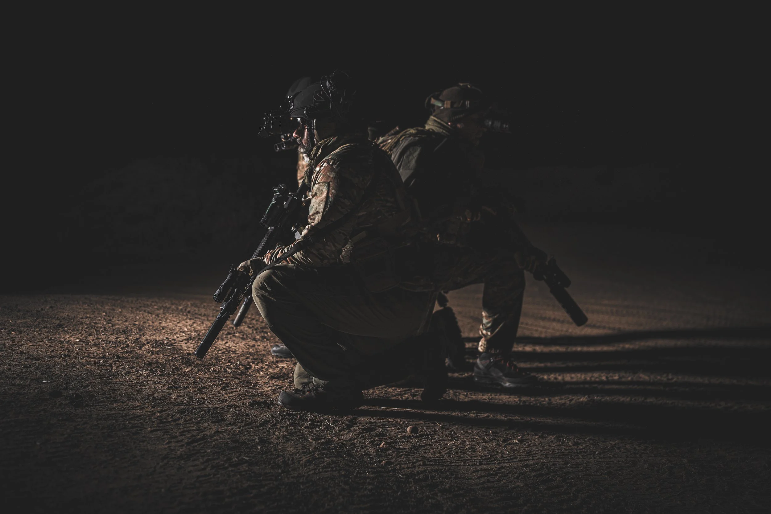 Two soldiers in camouflage uniforms and tactical gear are kneeling on the ground at night, aiming their rifles forward.
