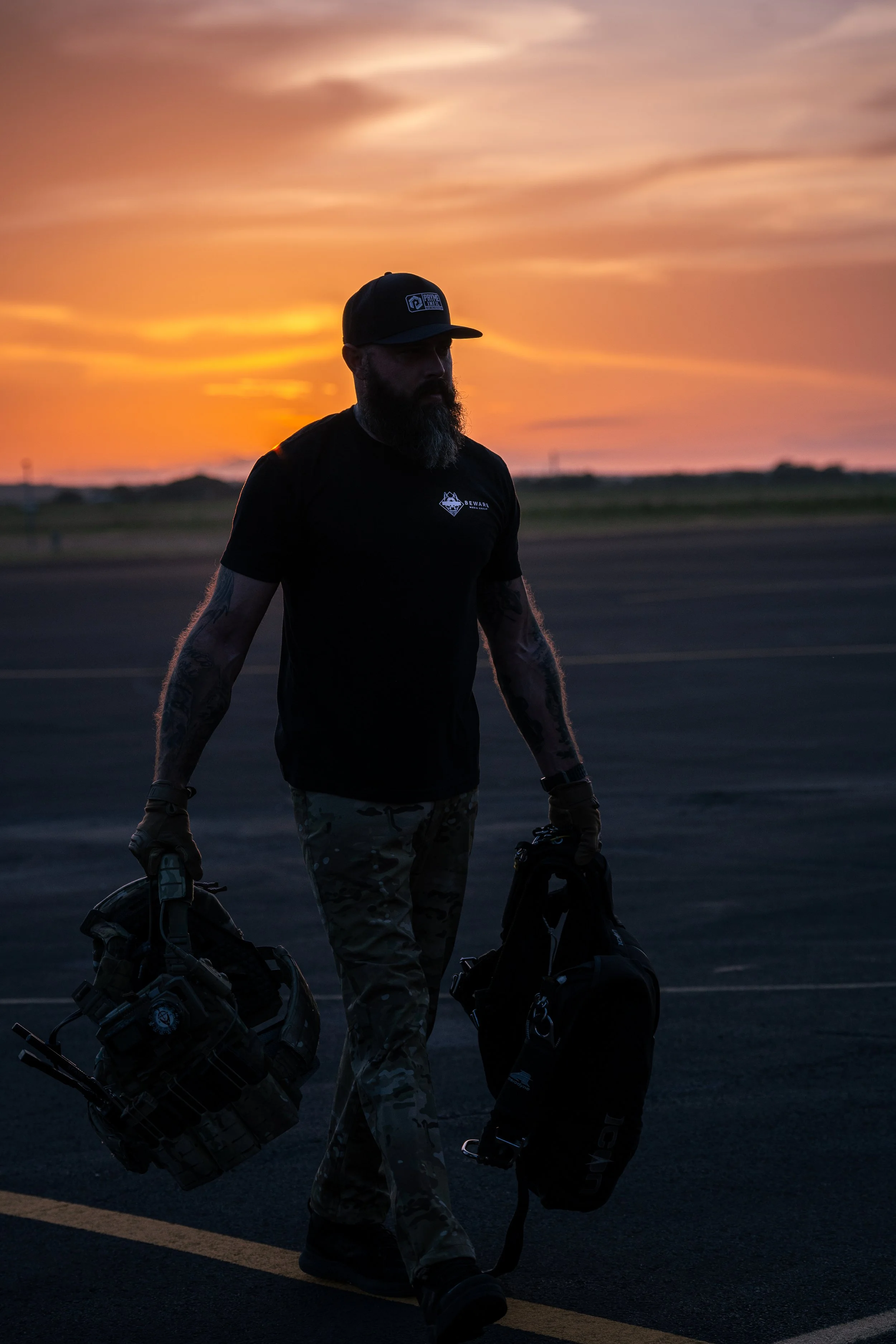 A man with a beard, tattoos, and a black cap walking on a tarmac at sunset, carrying two bags.