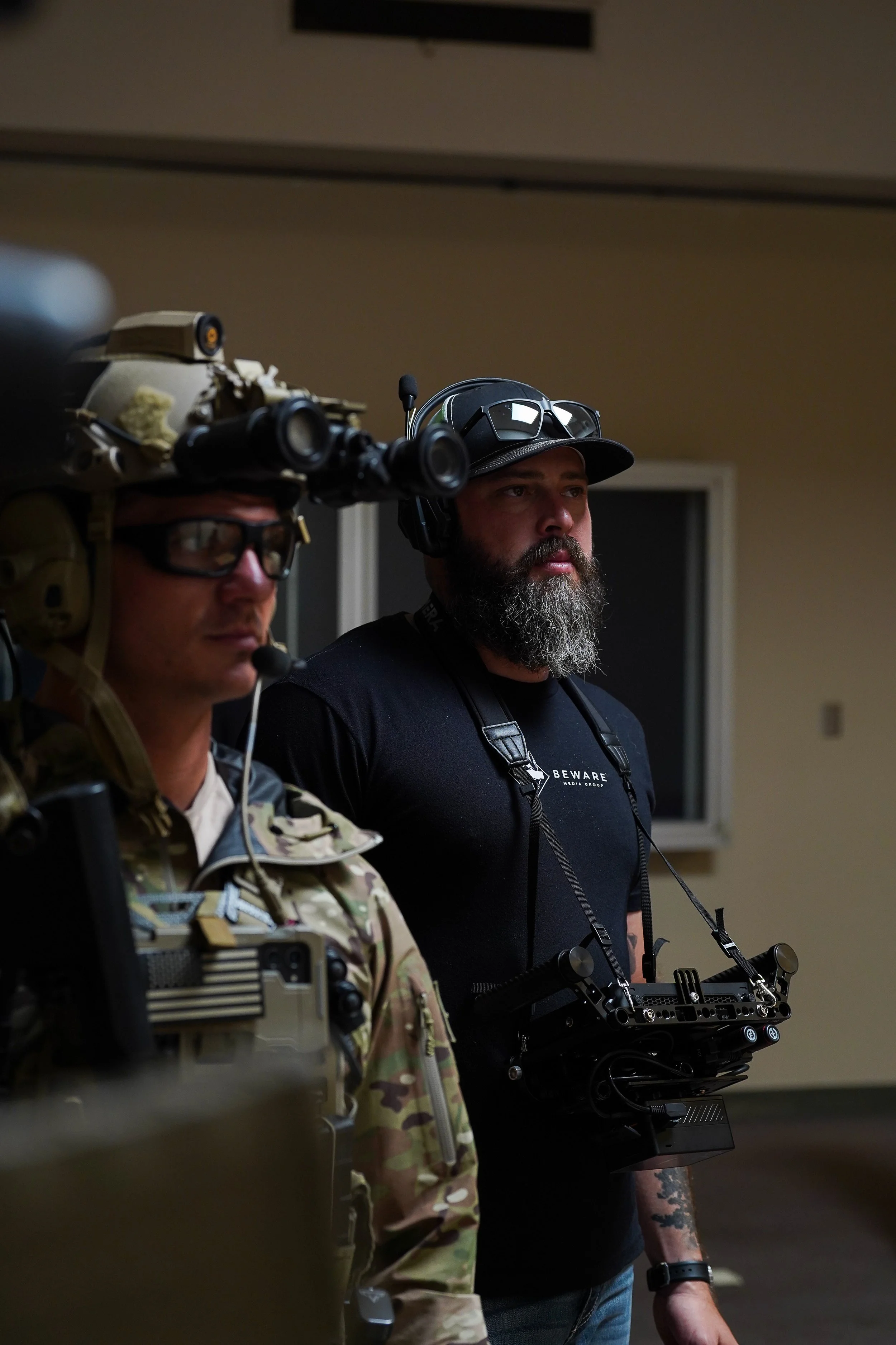 Two men standing indoors, one in military gear with a headset and night vision equipment, the other in casual clothing with a headset and a remote control device, both appear focused.