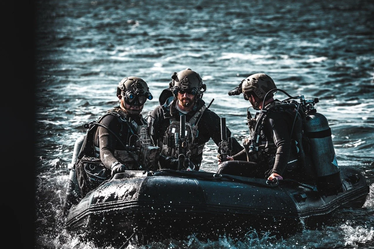 Three military personnel in tactical gear on a black inflatable boat in the water.