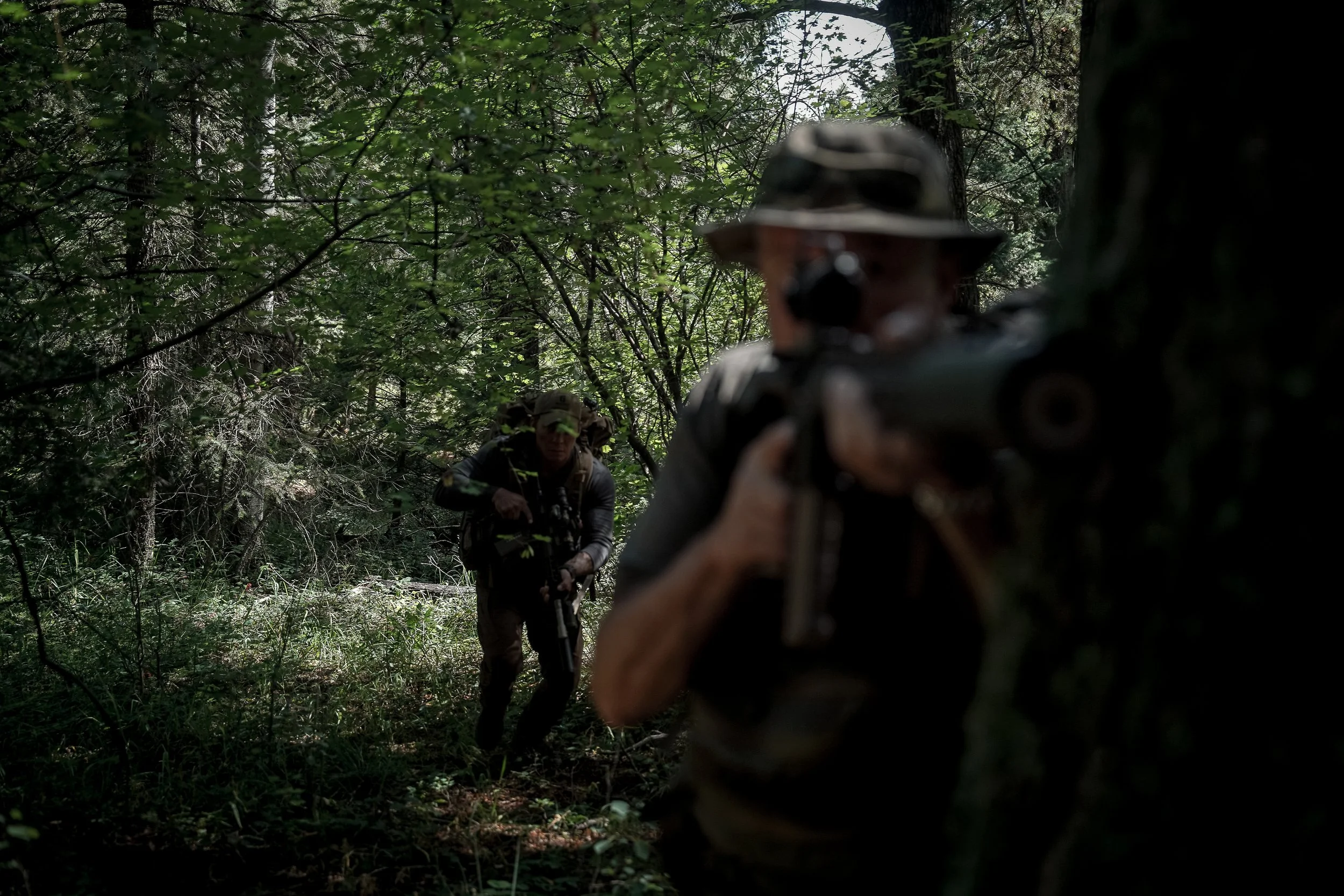 Two individuals in camouflage gear and wide-brim hats aiming rifles in a dense forest with green foliage.