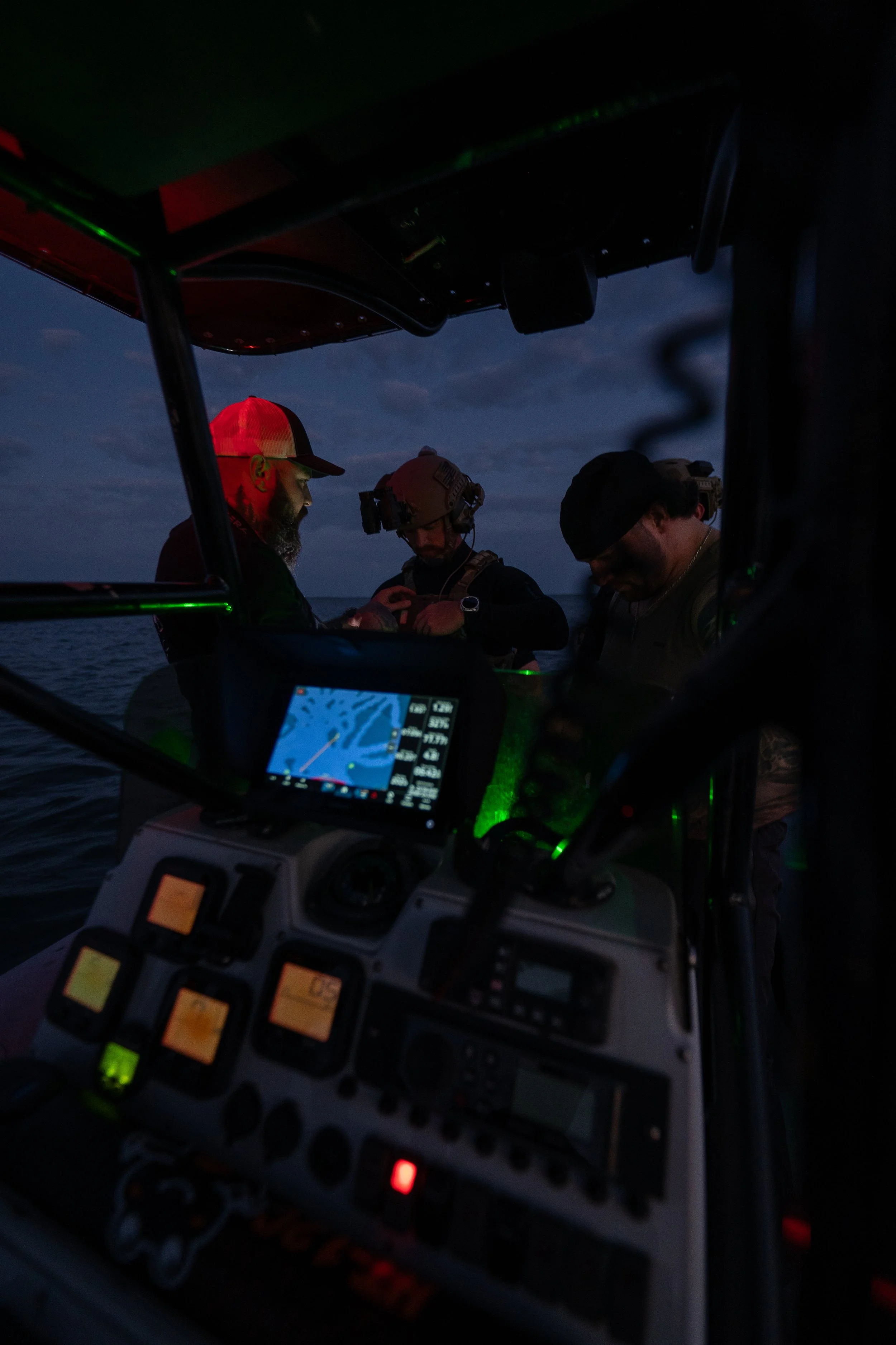 Three people in tactical gear gathered around equipment on a boat at dusk, illuminated by green and blue lights.