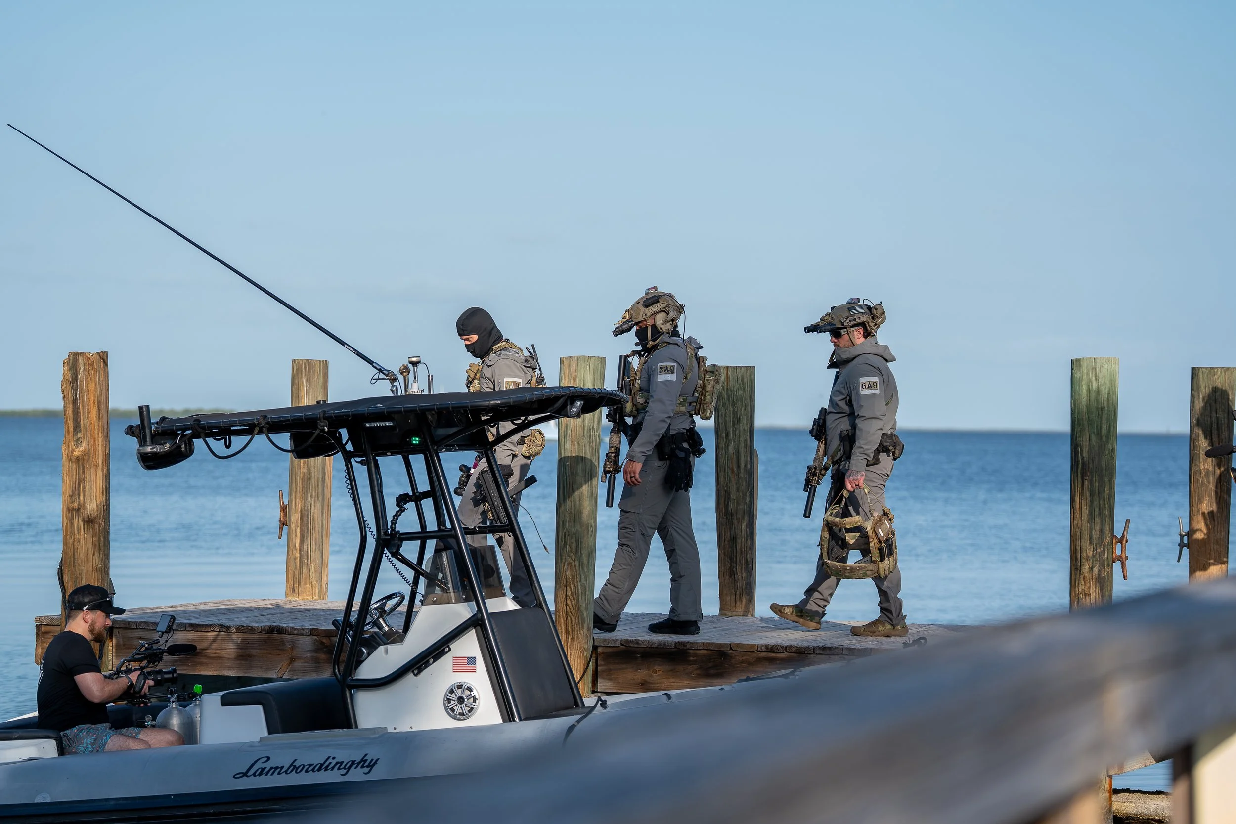 Three tactical police officers walking on a wooden dock beside a body of water, with a boat labeled 'Lamborghini' docked nearby, and a man operating a camera on the boat.