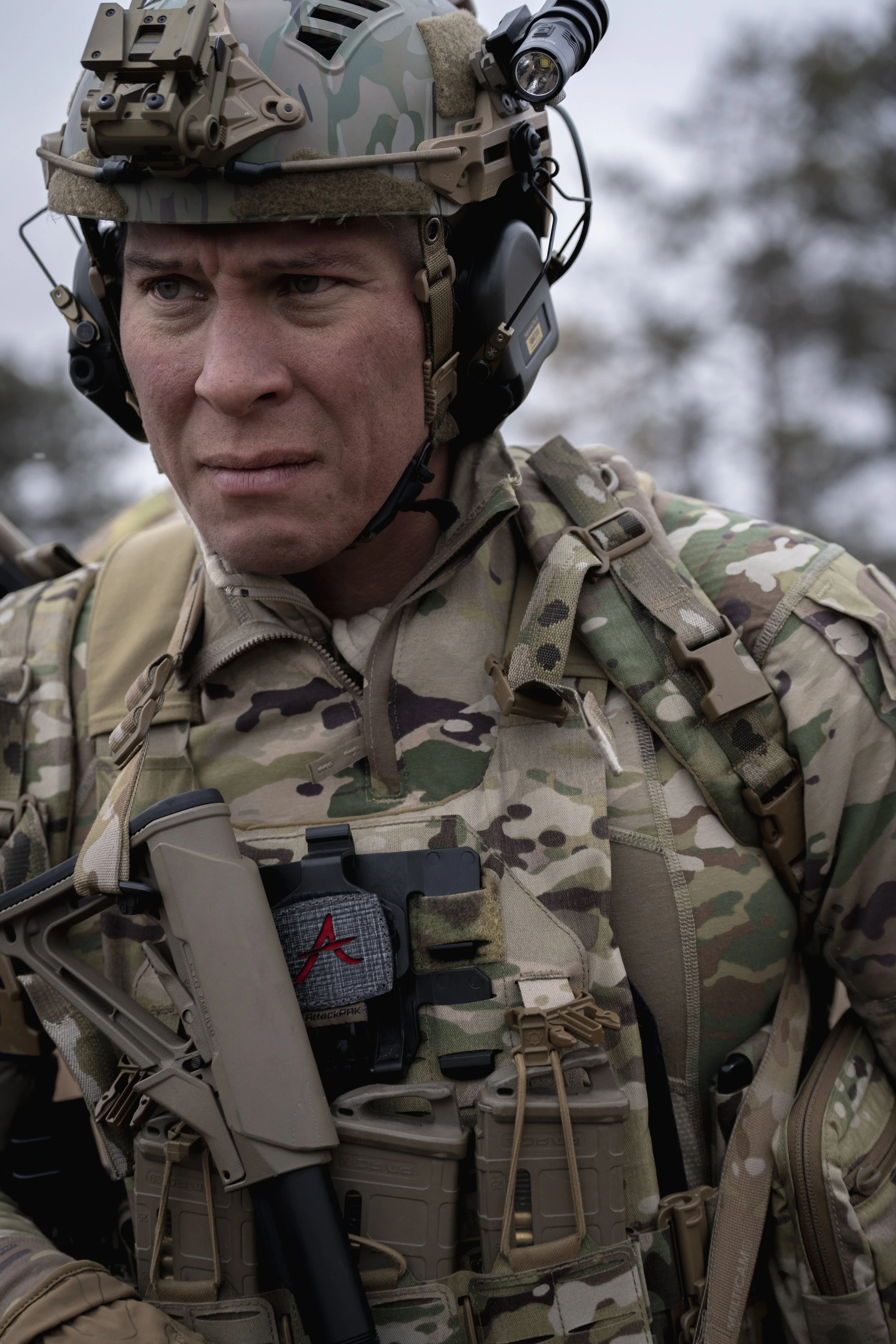 A male soldier dressed in camouflage uniform and tactical gear, including a helmet with mounted flashlight, and carrying a firearm, appears focused outdoors.