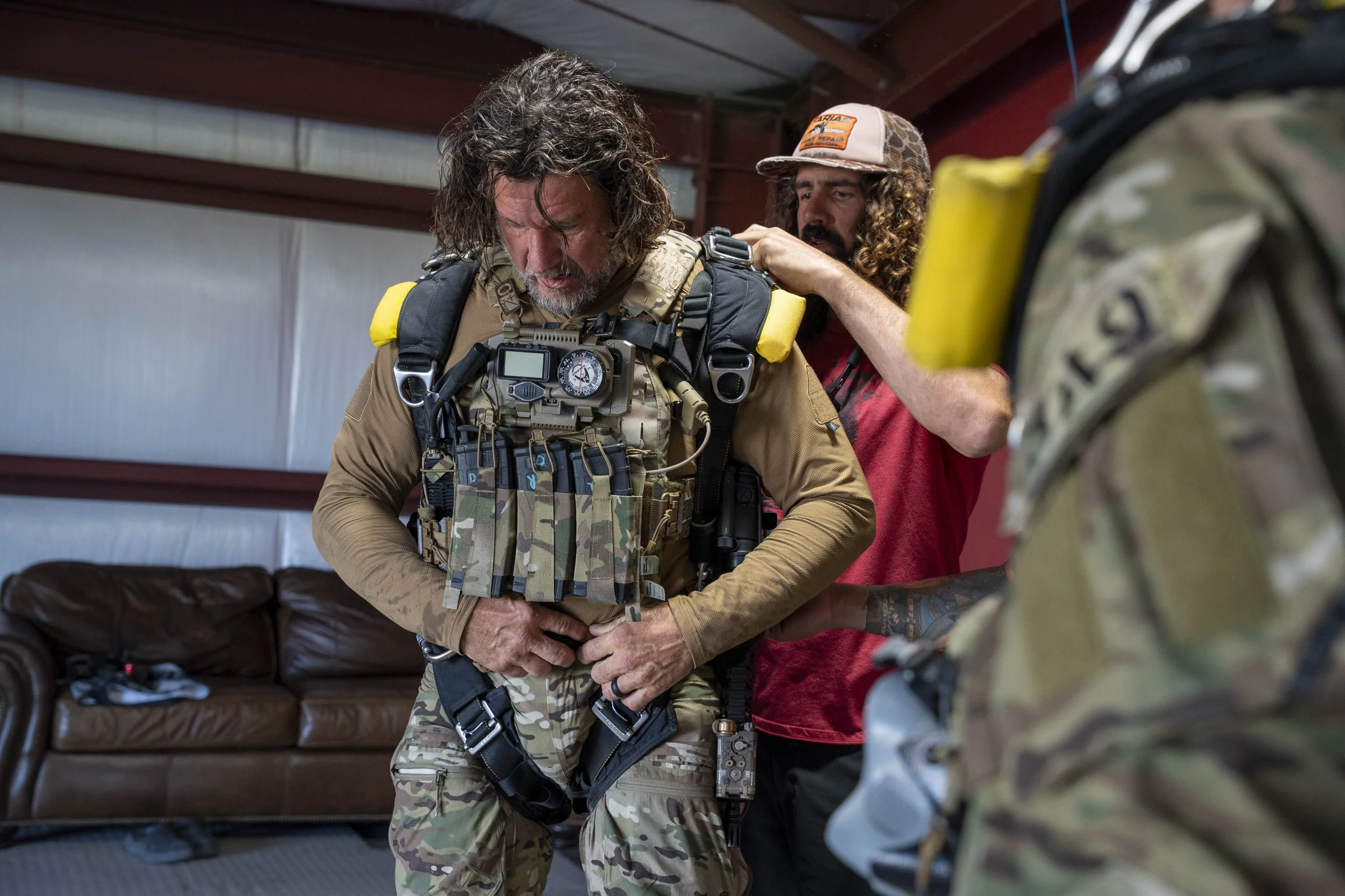 A man in military tactical gear adjusting his belt while another man helps him, inside a, indoor setting with a brown couch in the background.