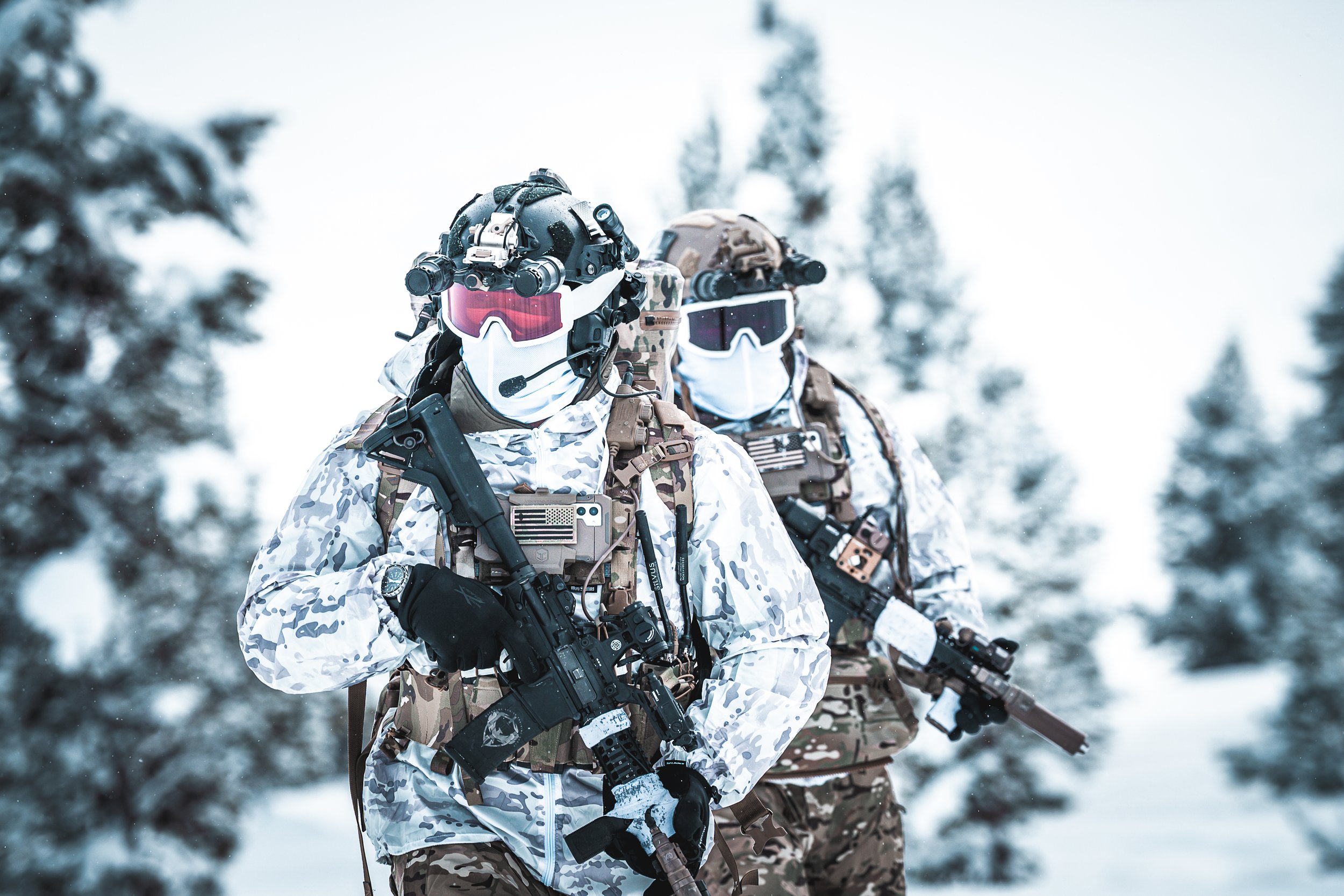 Two soldiers dressed in camouflage winter gear with tactical equipment, holding rifles, walking in a snowy forest.