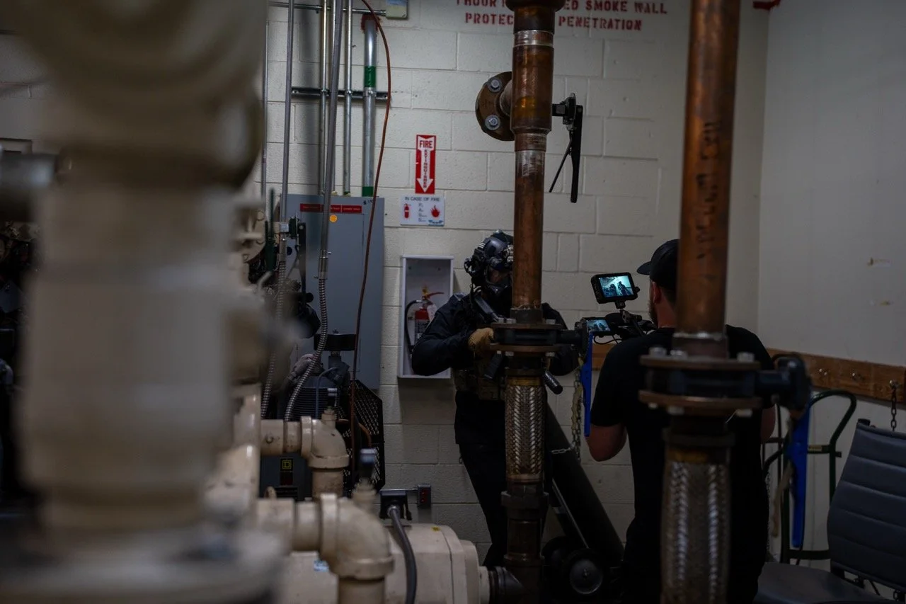 Two individuals conducting an inspection or recording in a mechanical room with pipes and equipment.