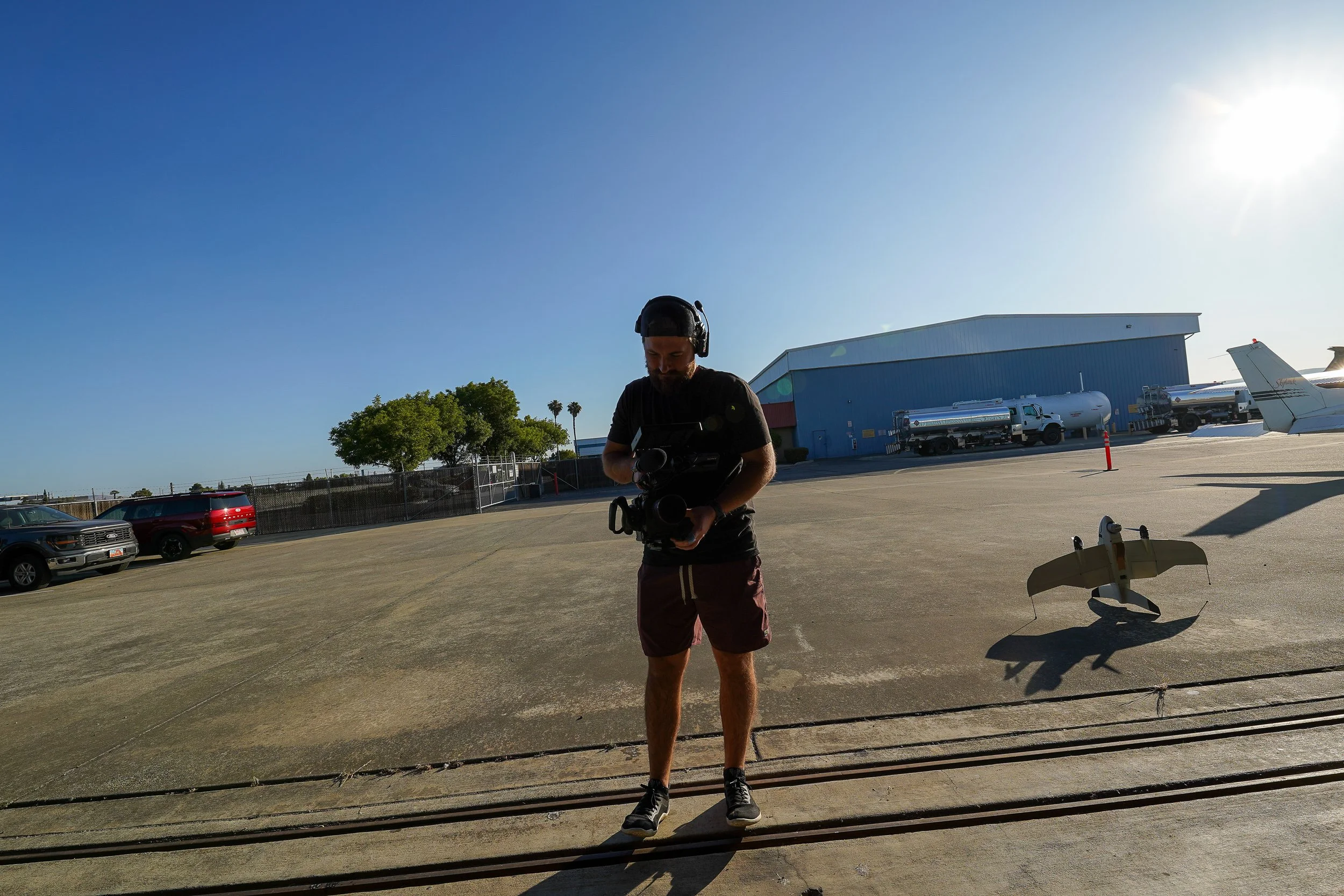 Man with headphones operating a video camera at an airport tarmac, with small airplane, fuel trucks, and airport hangar in the background, sunny weather with clear blue sky.