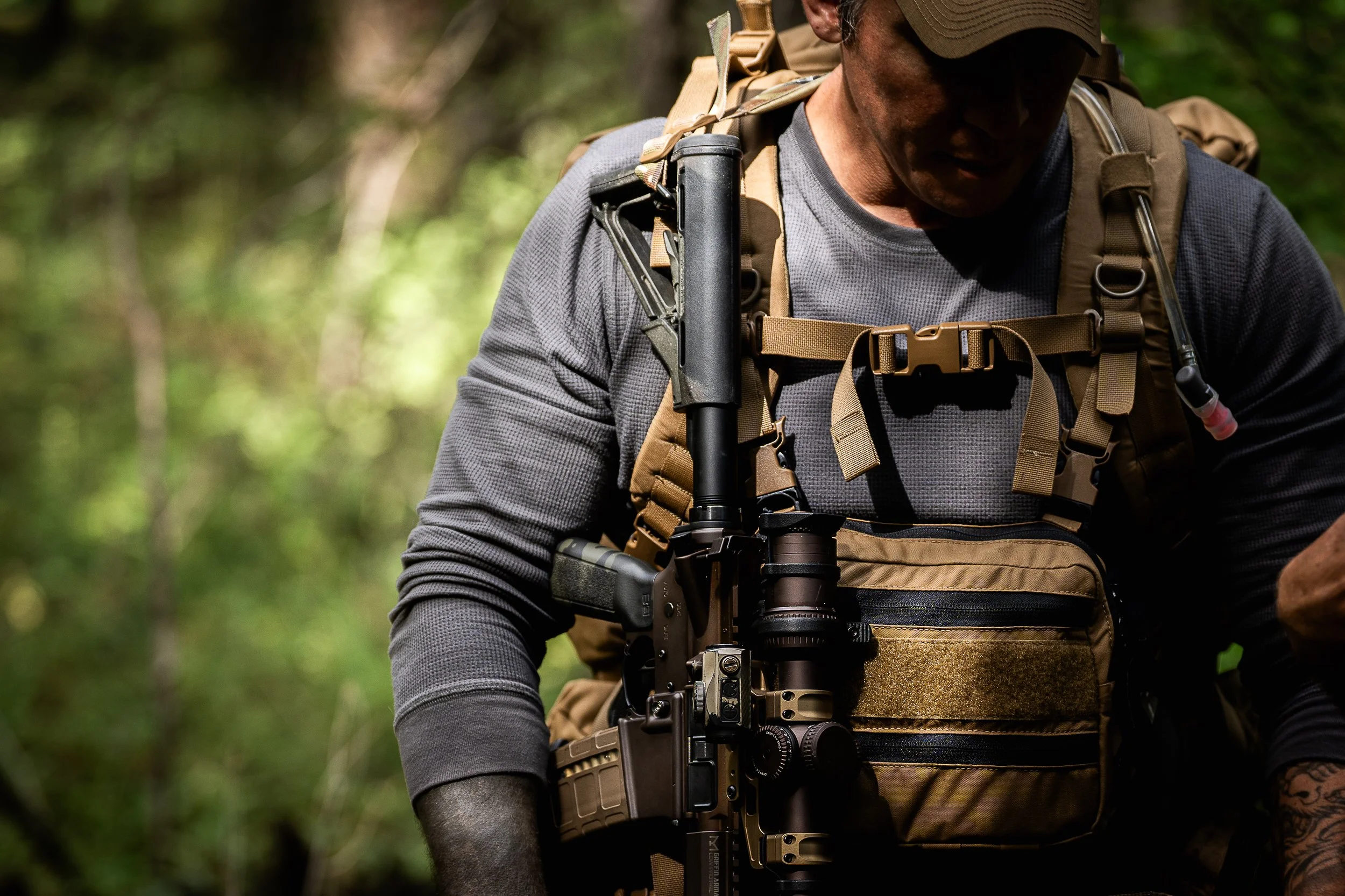 Close-up of a man in outdoor gear with tactical equipment, including a rifle, in a forest setting.