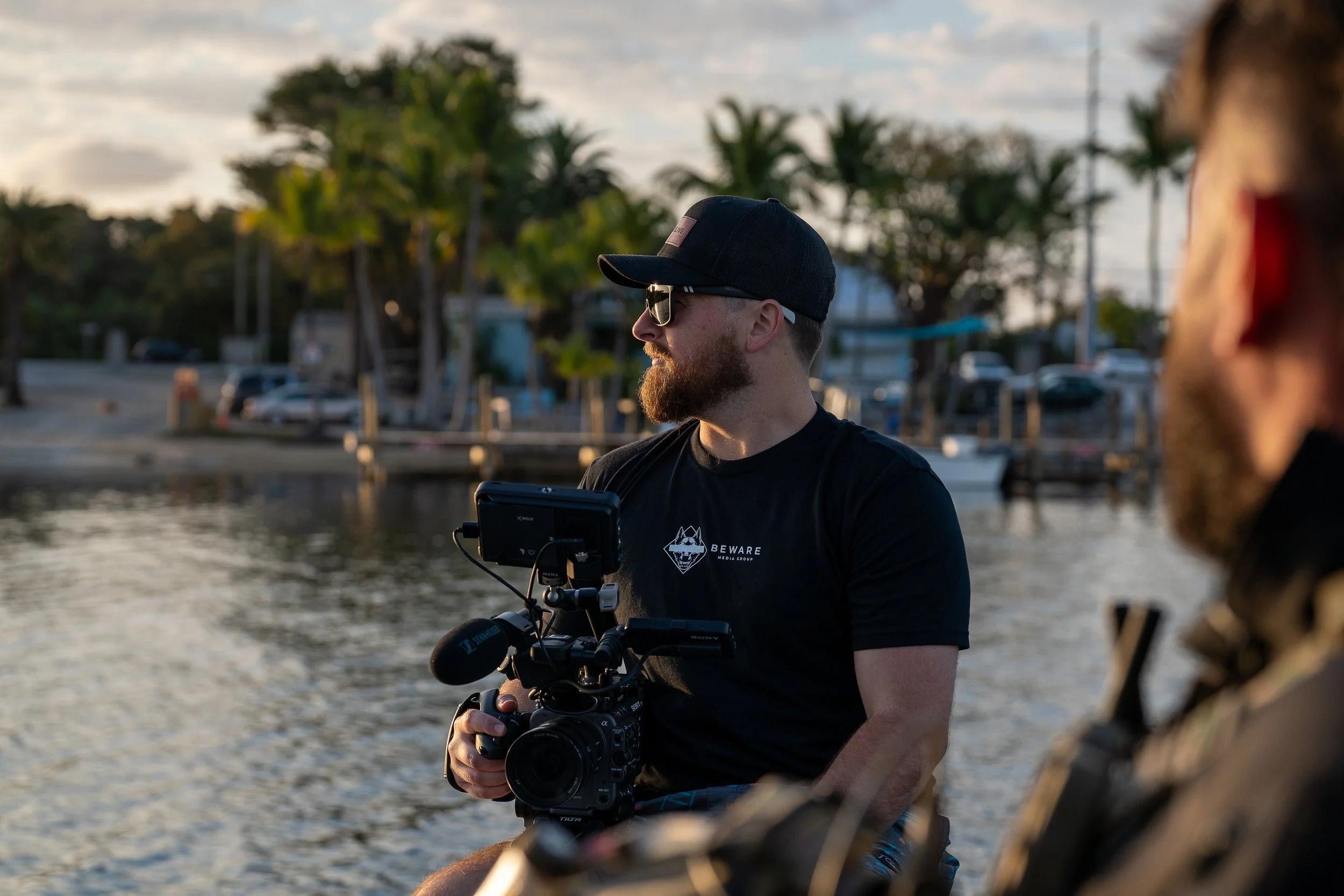 Two men with beards and casual clothing, one wearing sunglasses and a black cap, sit near a body of water with trees and boats in the background, during sunset.