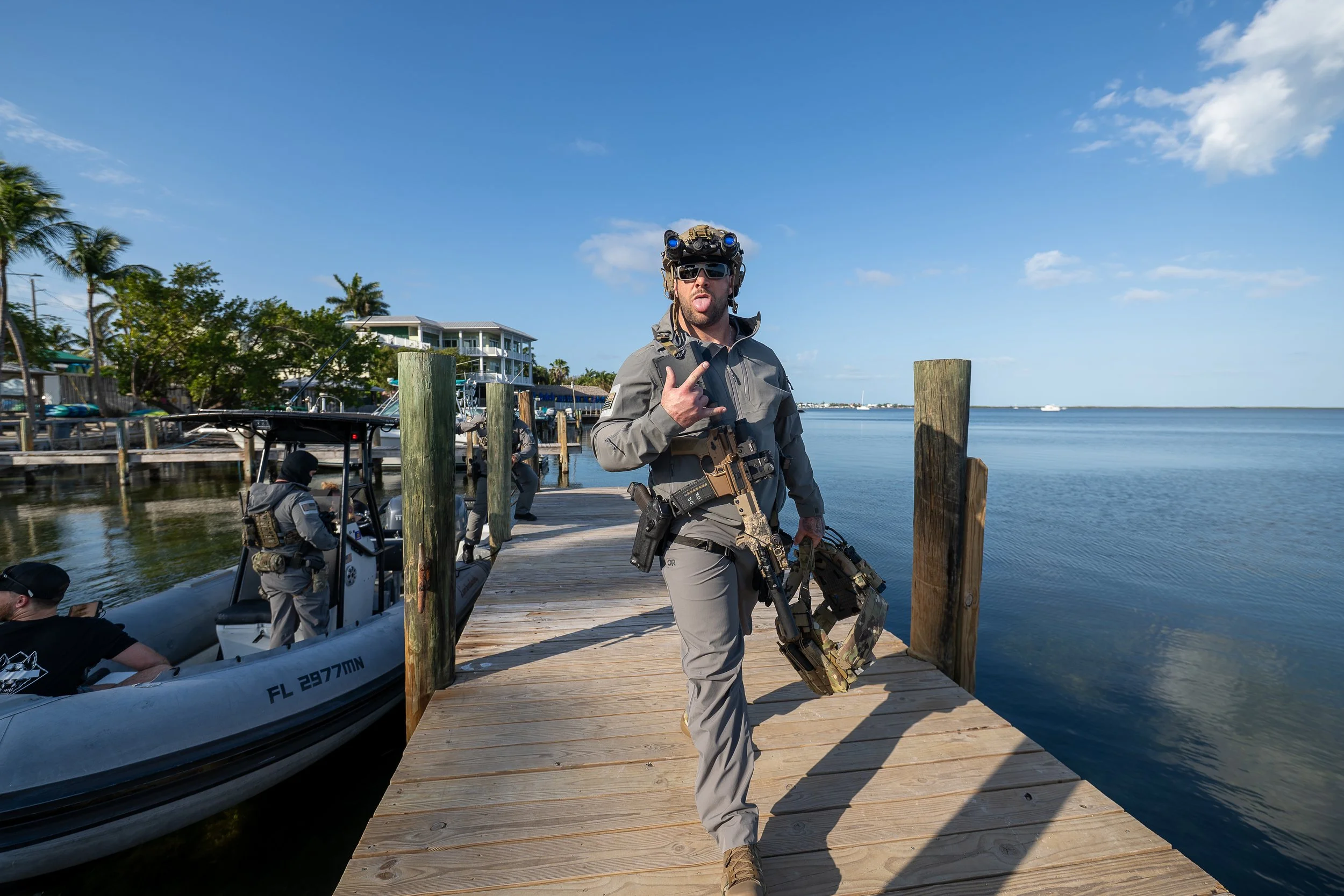 A man dressed in tactical gear, holding a firearm, walking on a wooden dock next to a boat by the water, making a gesture with his hand, under a clear blue sky.
