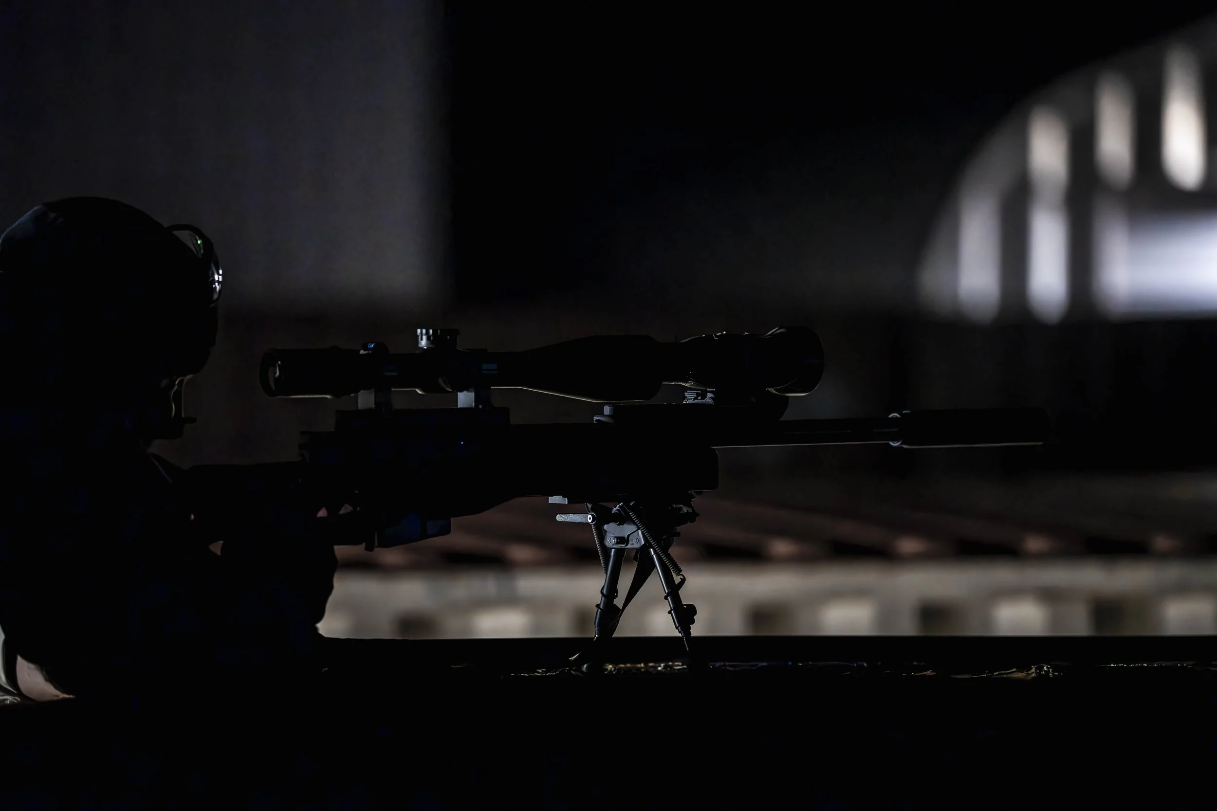A person wearing a helmet aiming a scoped rifle in a dark environment, with a faint light source illuminating part of the background.