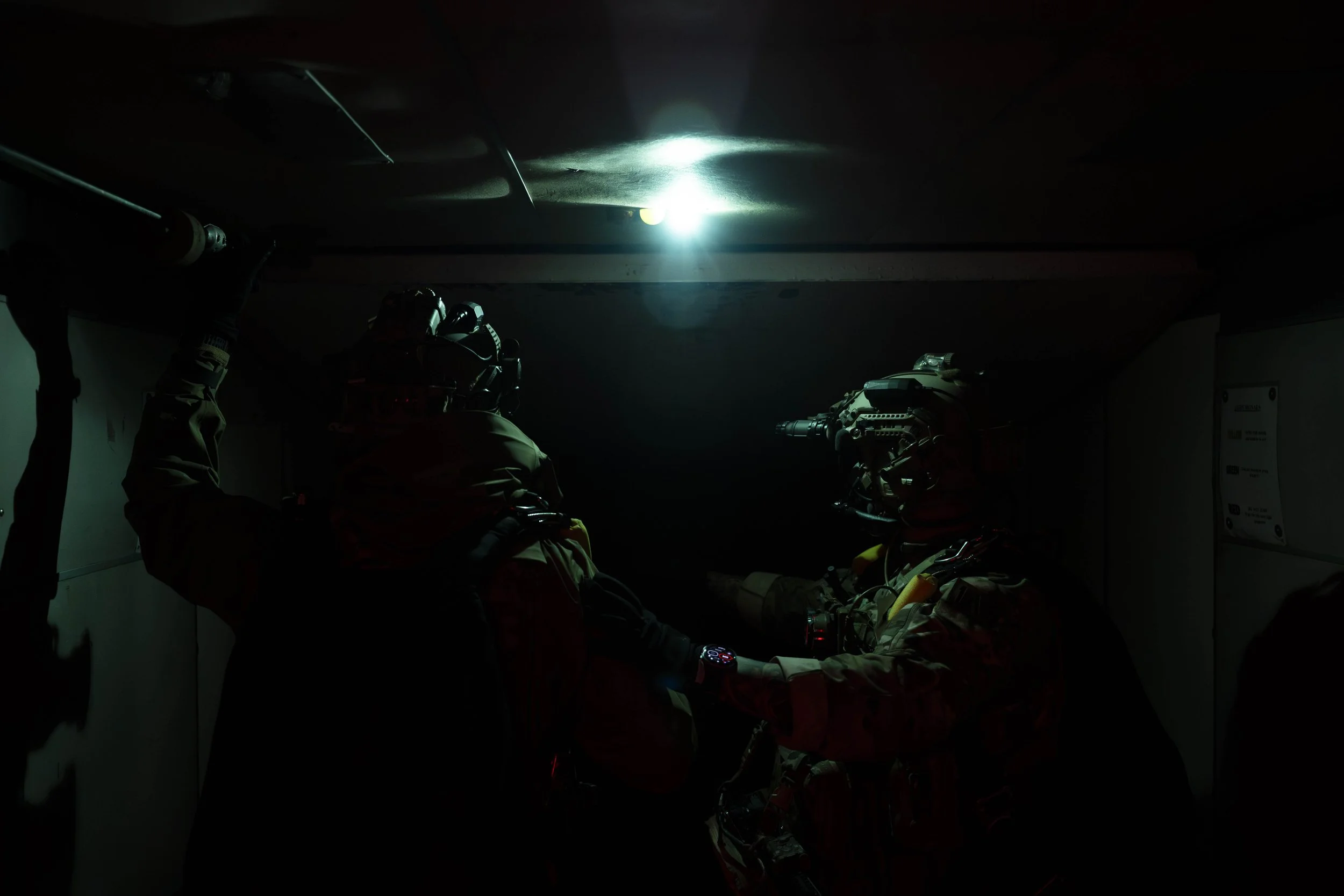 Three soldiers in tactical gear exchanging a hand shake in a dark room with a ceiling light overhead.
