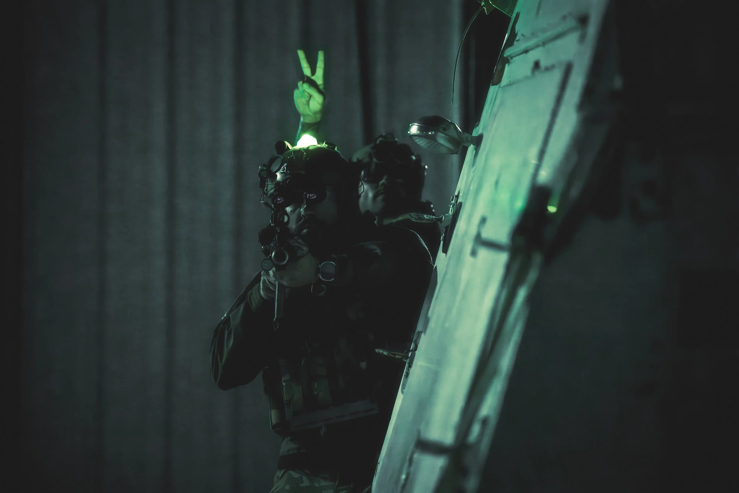 A person dressed in tactical gear, wearing a helmet with lights, is climbing a ladder in a dark environment. The person is making a peace sign with their hand, which is illuminated by a green light.