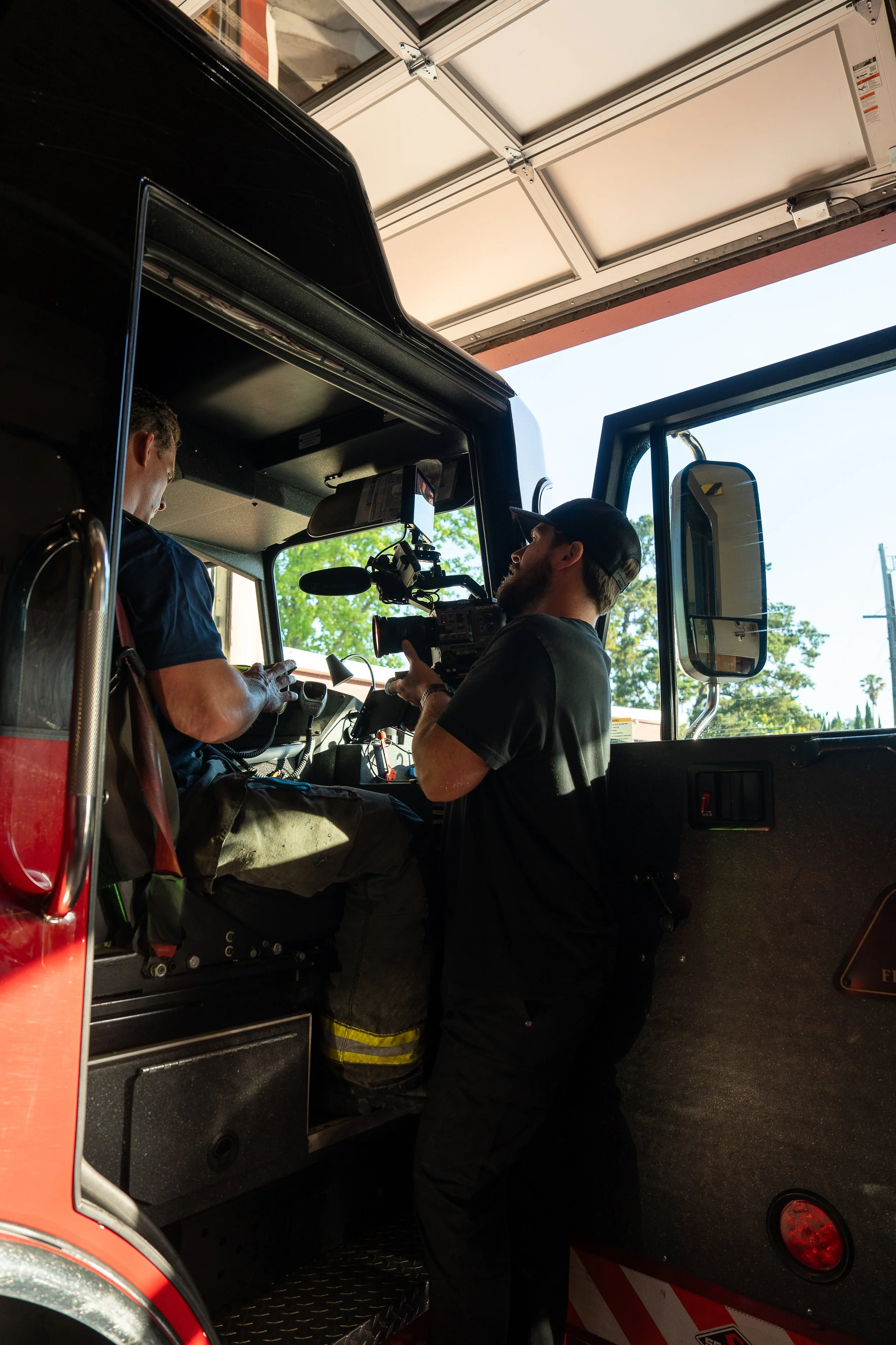 Crew filming inside a fire truck with the driver sitting in the driver's seat and a cameraman operating a professional camera.
