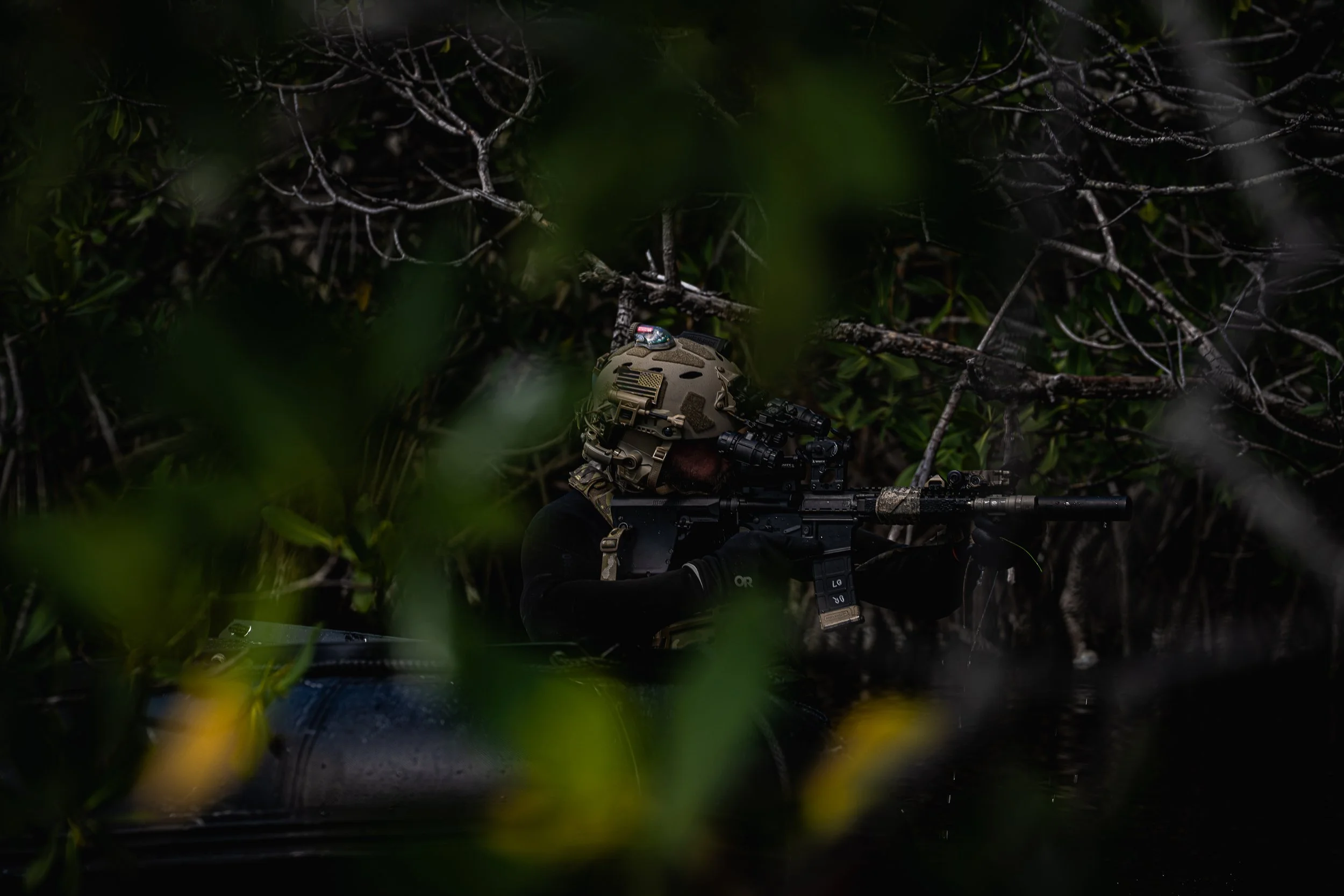 A soldier in camouflage gear aiming a sniper rifle through dense green foliage in a jungle setting.