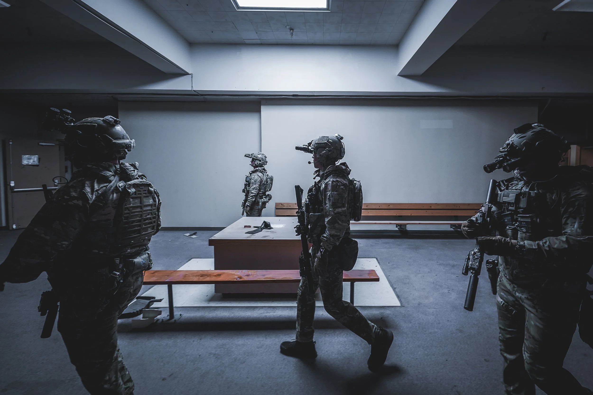 Four soldiers in camouflage uniforms and tactical gear standing and walking in an indoor setting, possibly a training or briefing room, with a wooden bench and a table in the background.