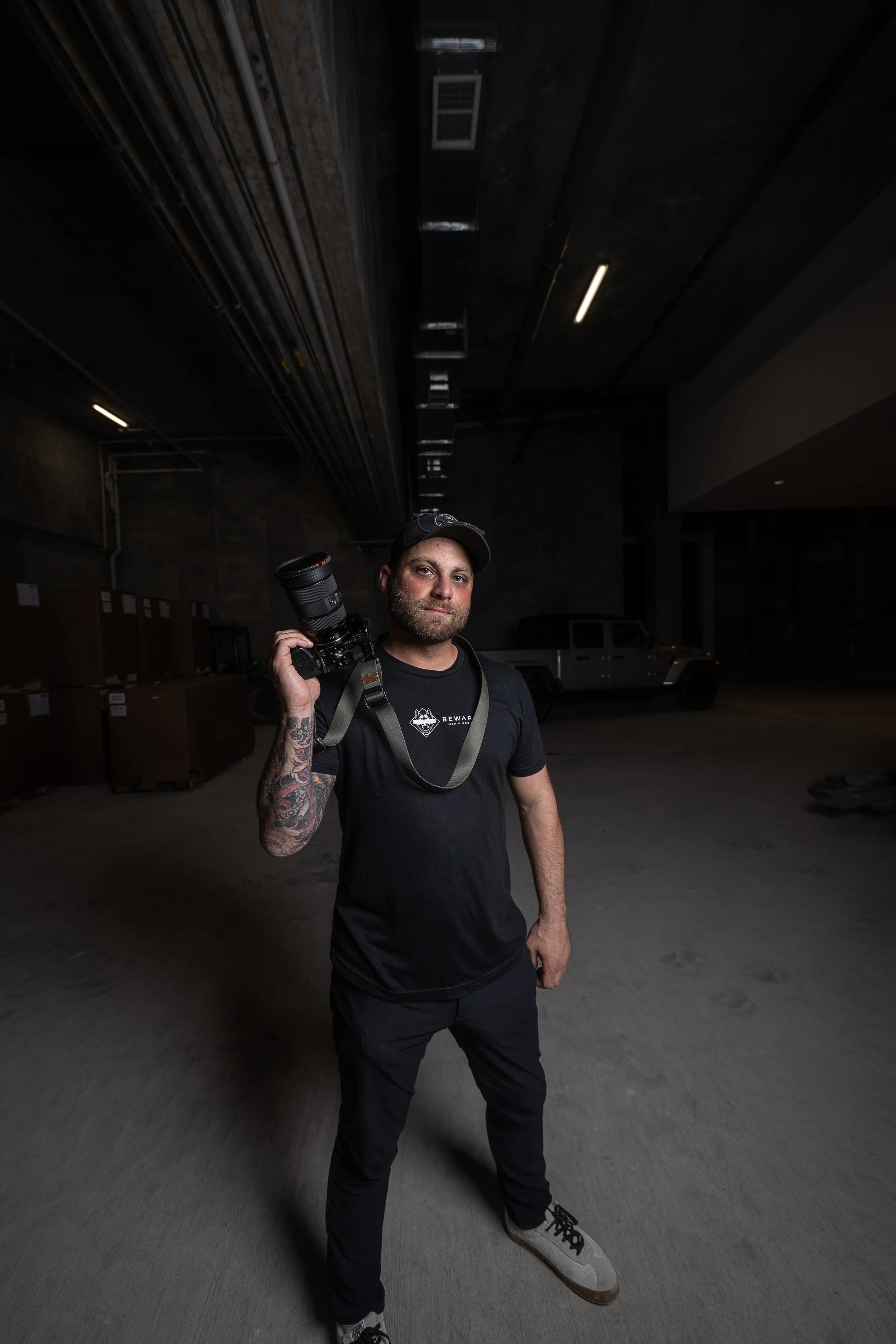 A man standing in a dimly lit parking garage, holding a camera with a large lens over his shoulder, wearing a black t-shirt, black pants, a black cap, and white sneakers.