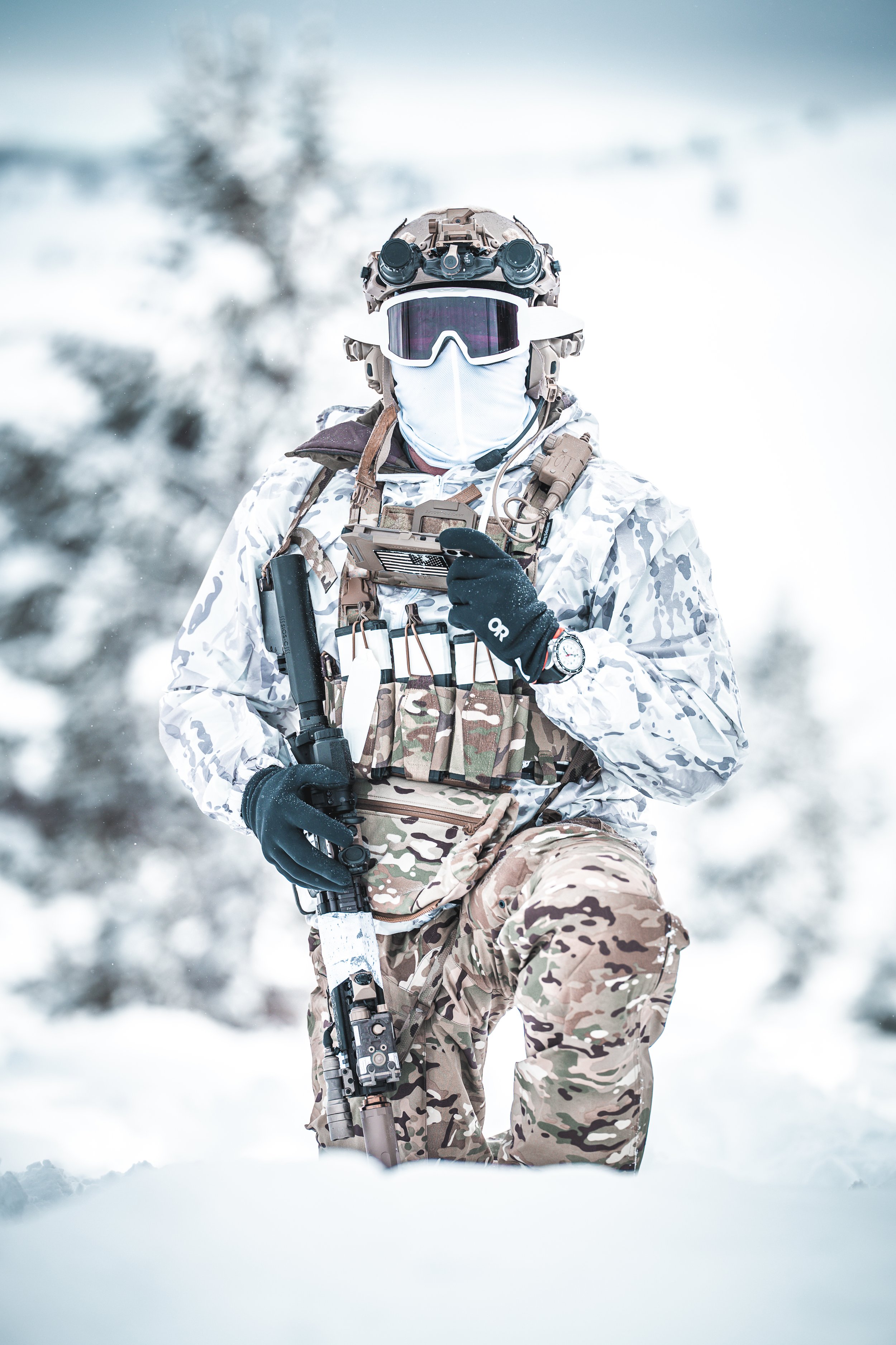 Soldier in camouflage winter gear with tactical vest, helmet with goggles, face mask, gloves, holding a rifle, in snowy outdoor environment.