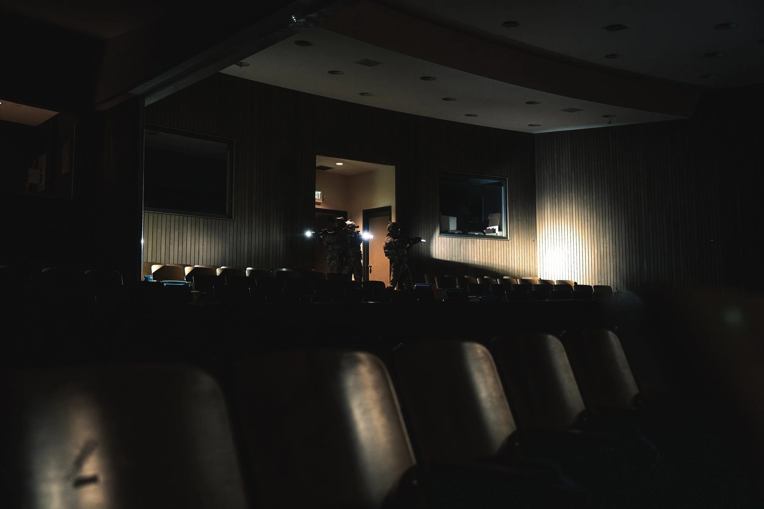 Two armed soldiers in tactical gear standing in a dimly lit auditorium with empty seats, near an open door with brightness coming through.