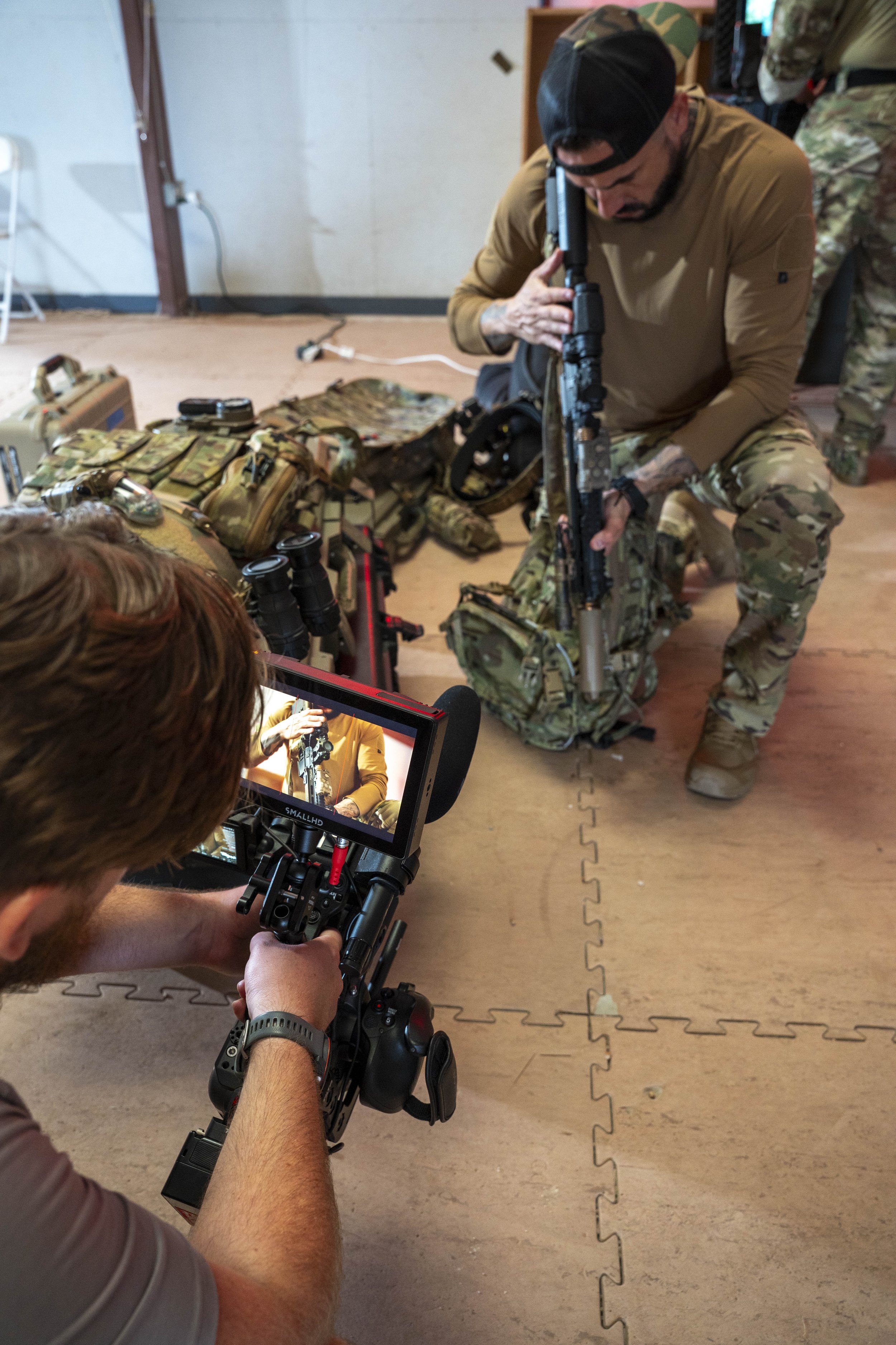 A man dressed in military camouflage gear is kneeling on the floor, holding a rifle in a shooting stance. A filmmaker or photographer with a camera mounted on a stabilizer is filming him. There are tactical gear and equipment on the floor around them