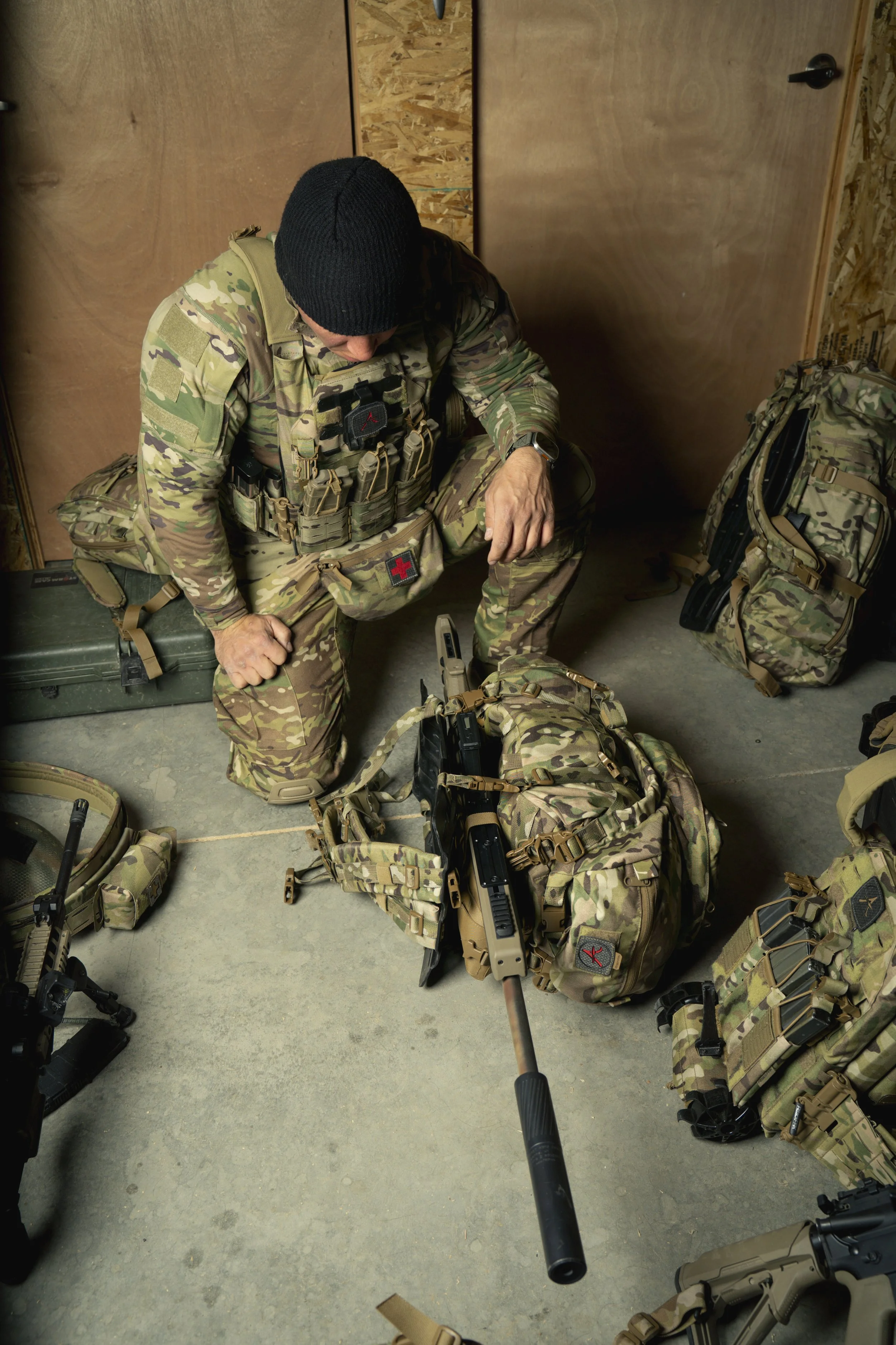 A soldier in camouflage gear and a black beanie kneeling on the floor, surrounded by military equipment, including rifles, backpacks, and tactical gear.