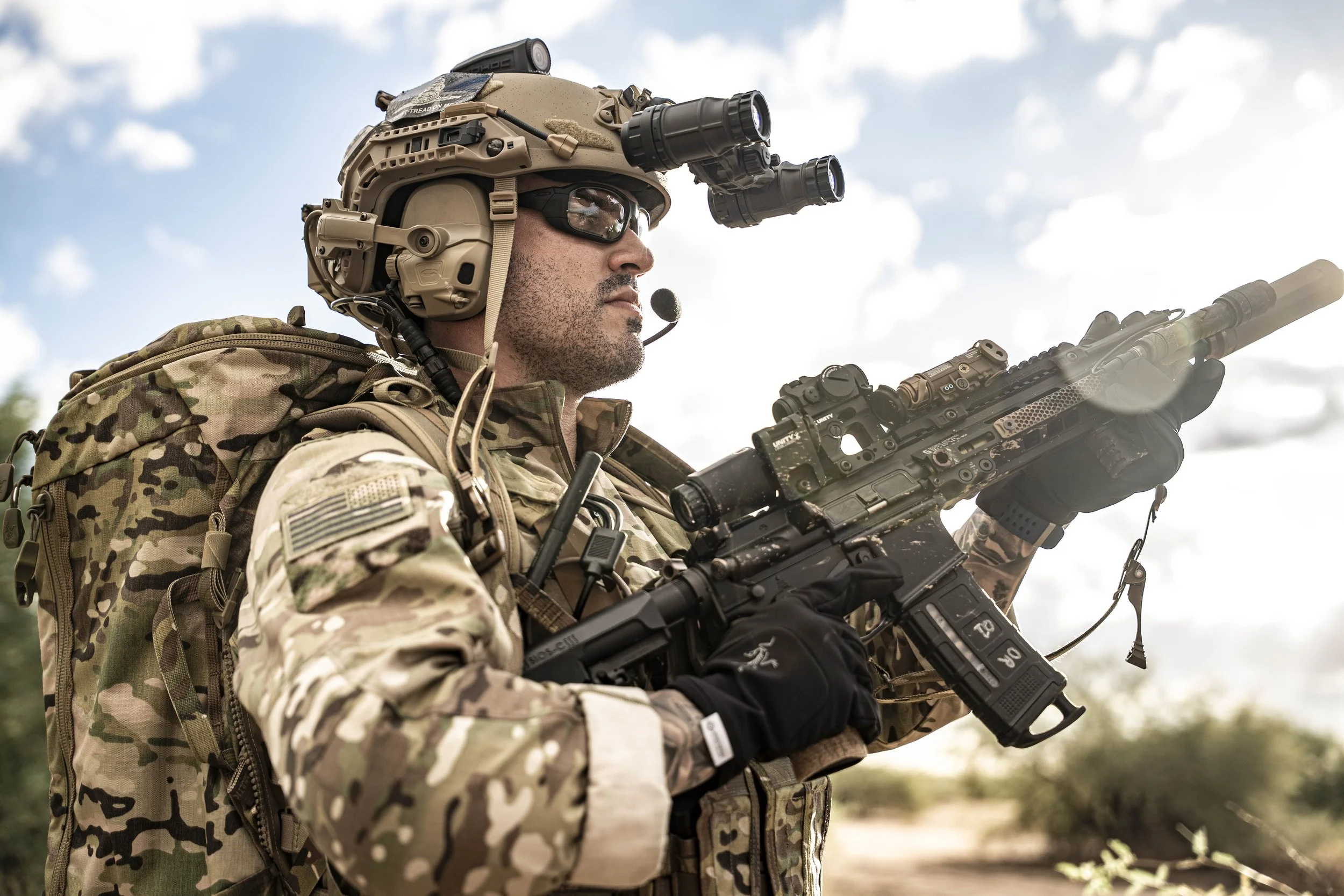 A soldier in camouflage gear aiming an assault rifle outdoors with a cloudy sky in the background.