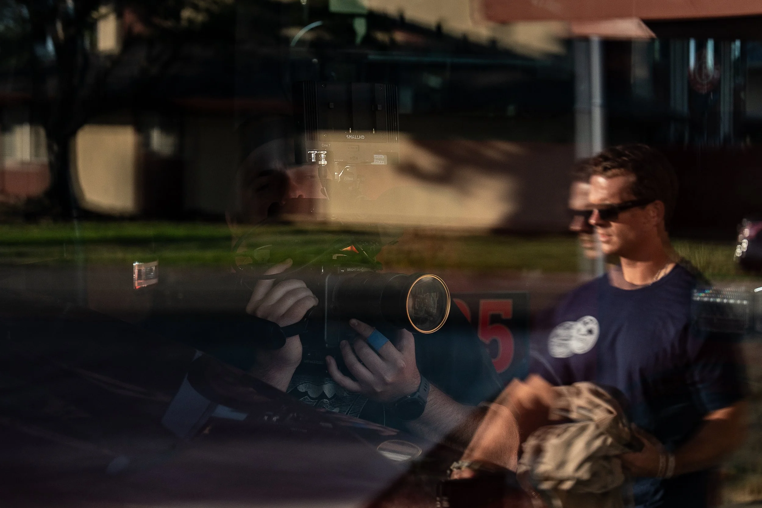 Reflection of a person taking a photograph with a camera, seen through a window, with a man standing outside wearing sunglasses in the background.