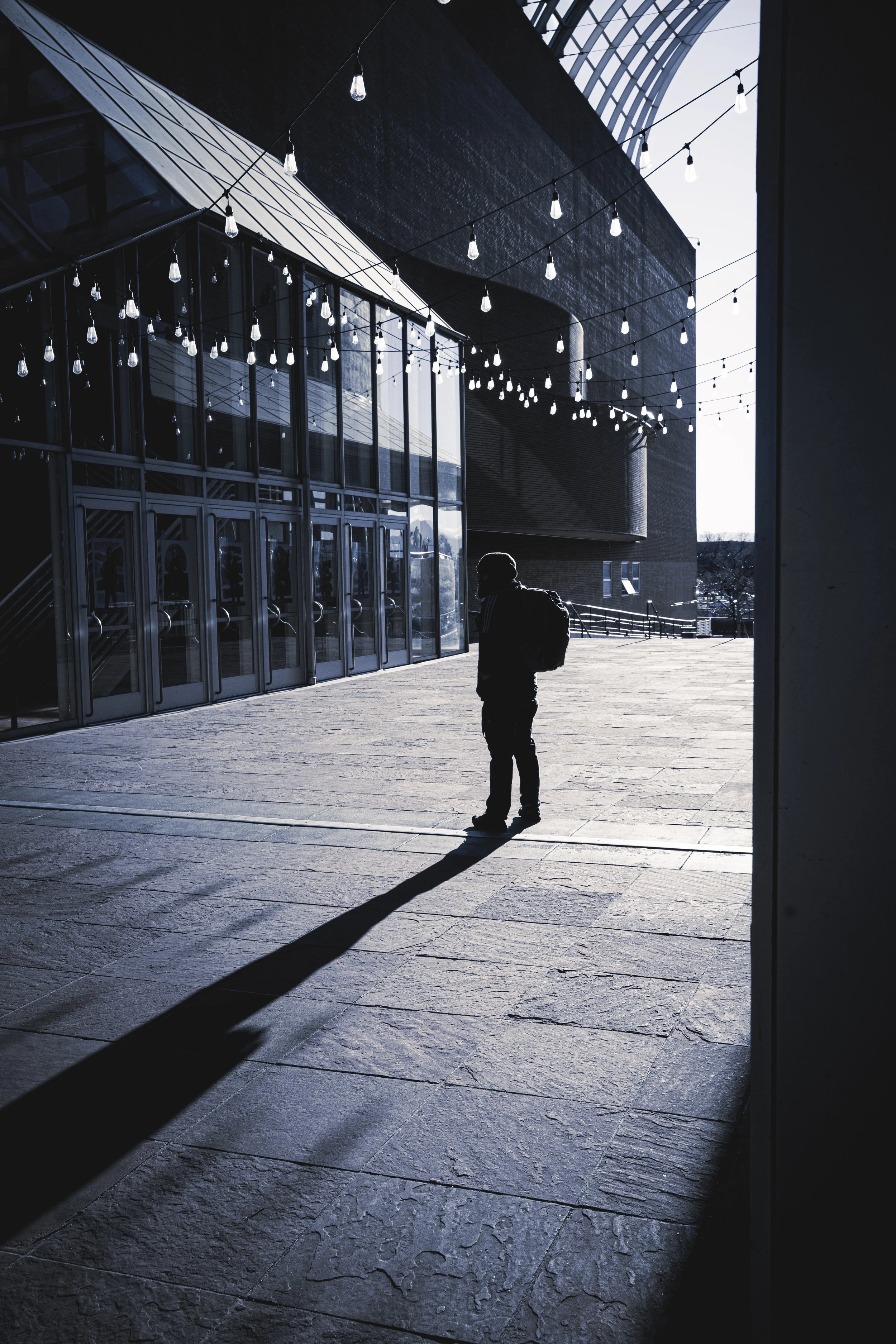Silhouette of a person with a backpack standing outside near a modern building with glass doors and string lights, cast in shadows during daylight.
