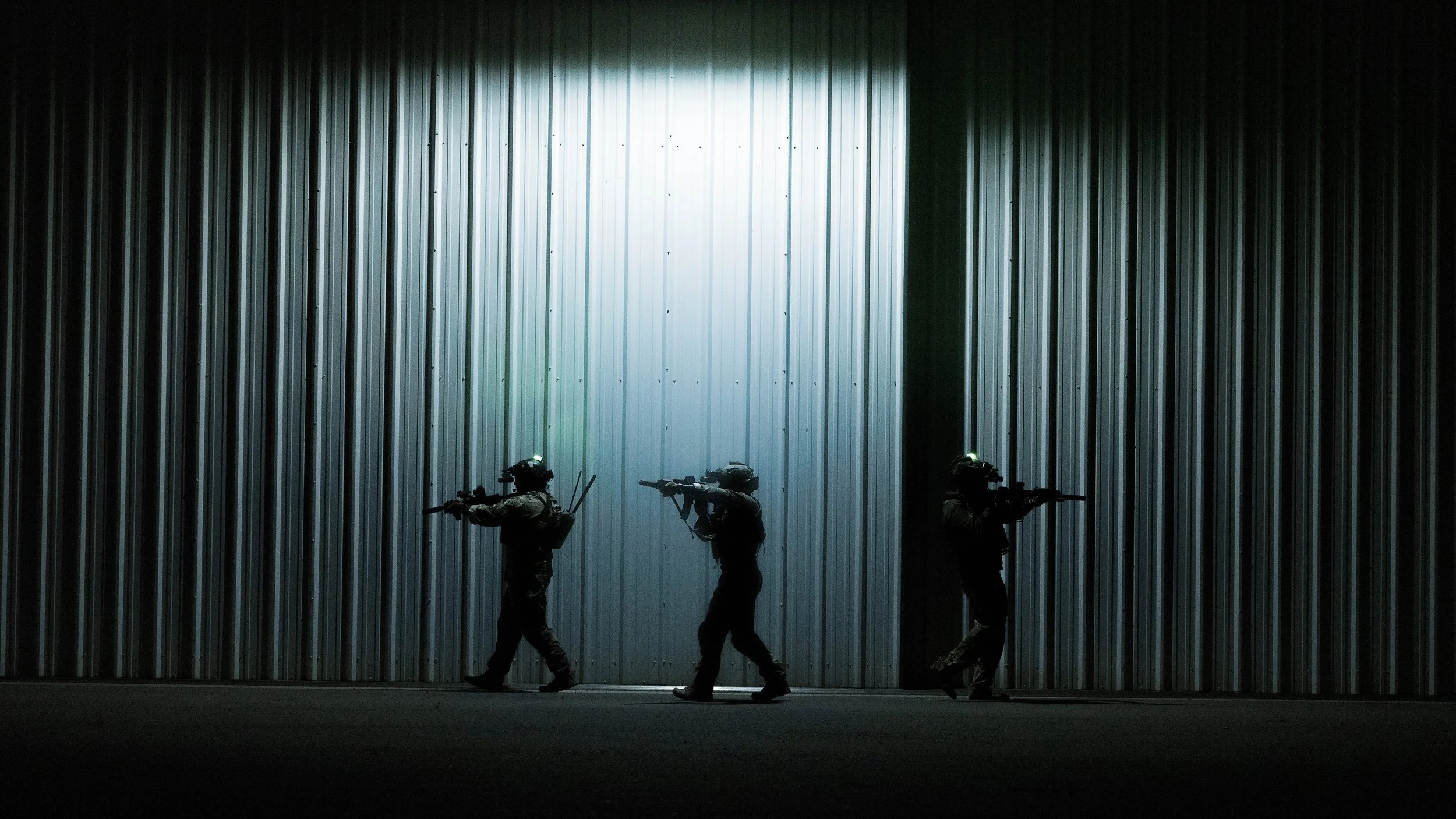 Three soldiers in tactical gear walking in front of a large, illuminated metal wall.