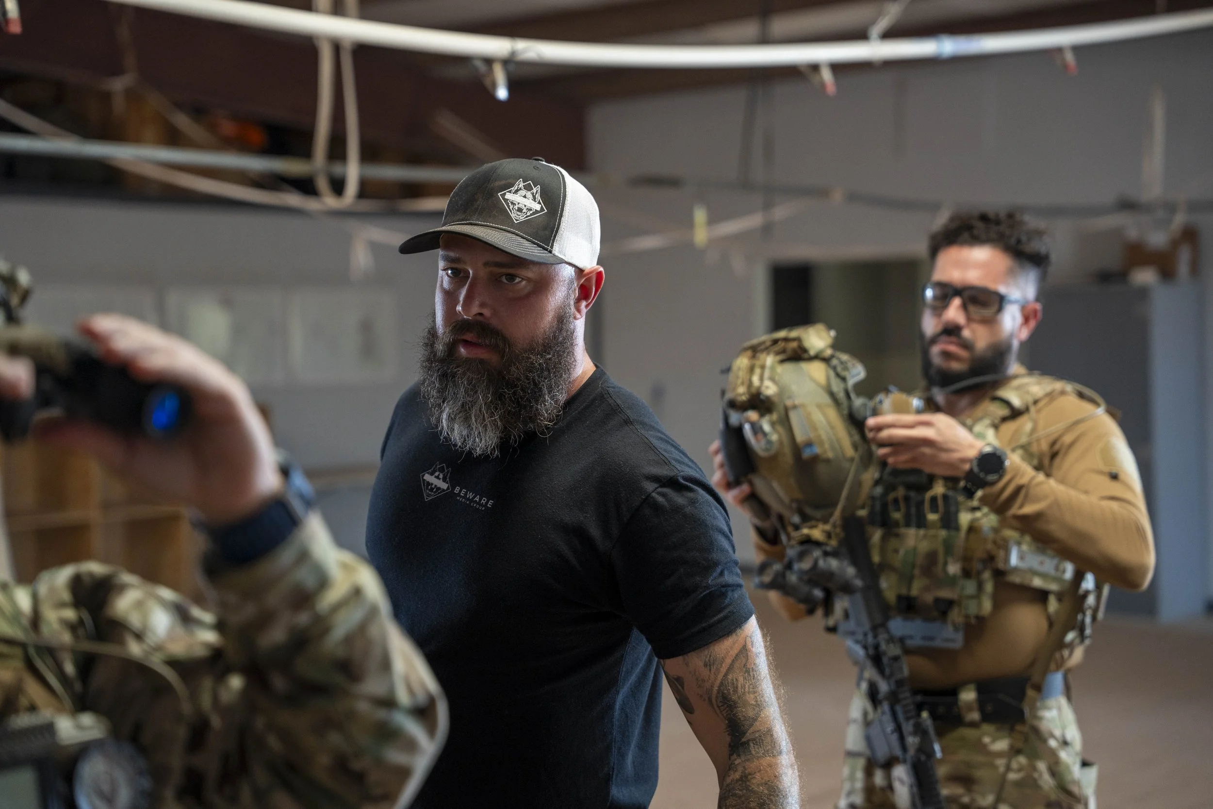 Two men in military gear and tactical clothing in a training or briefing room, with one holding a rifle and the other adjusting equipment.