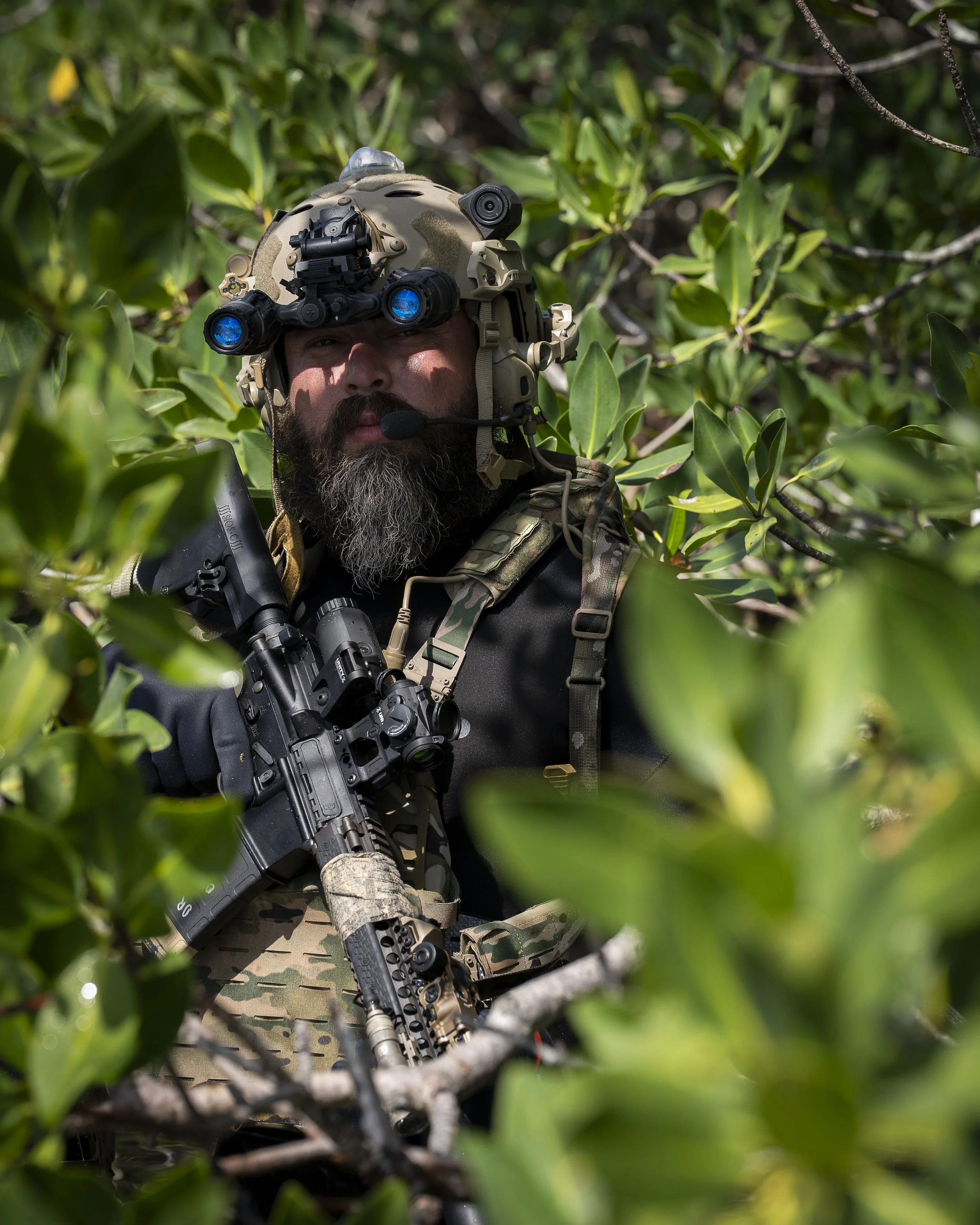 A military personnel in camouflage gear, wearing a helmet with night vision goggles, holding a rifle, and surrounded by dense green foliage.