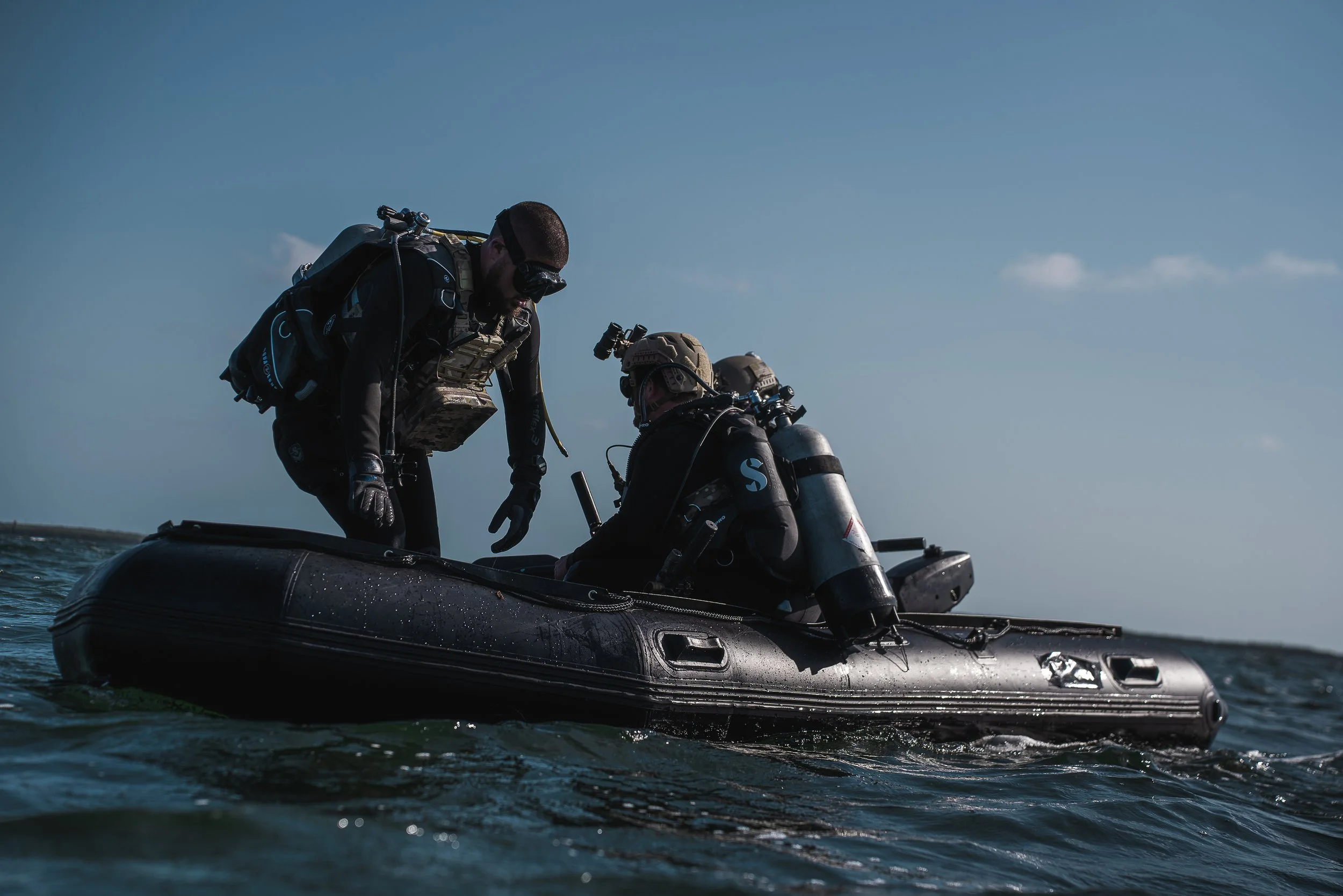 Two divers in wetsuits and diving gear preparing on a small inflatable boat in open water.