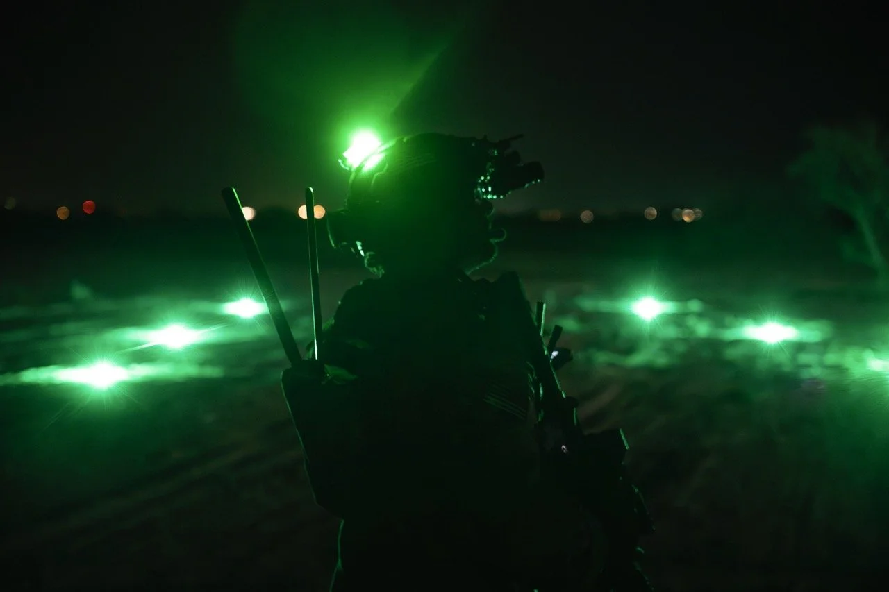 A soldier wearing night vision gear and a helmet with a mounted headlamp, standing in a dark outdoor setting illuminated by green lights.