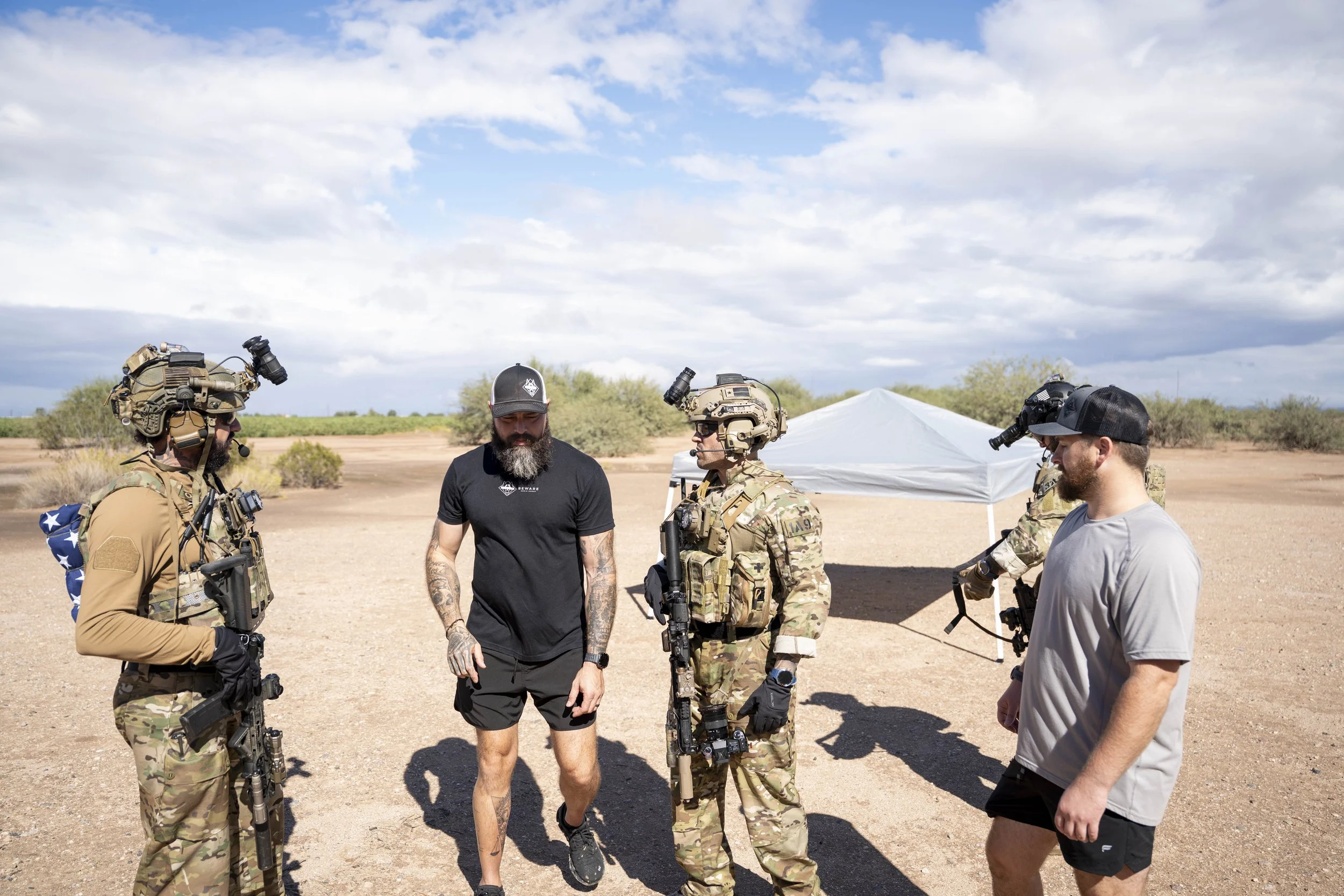 Group of soldiers and civilians standing outdoors in a desert landscape with a tent and scattered bushes.