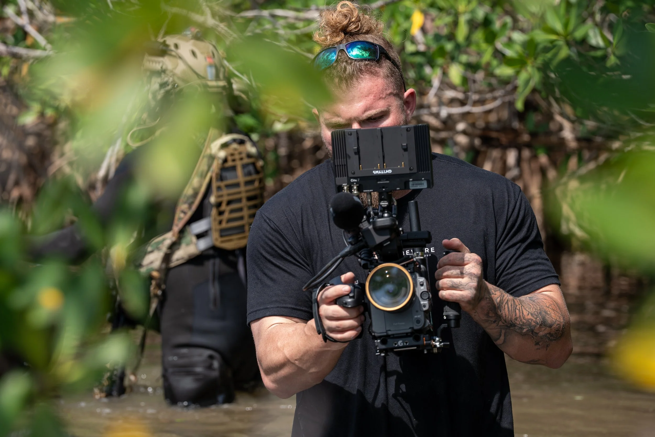 A man with tattoos on his arms holding a professional camera with a mounted monitor, taking photos or filming in a swampy area with green foliage and another person in the background wearing a helmet and outdoor gear.