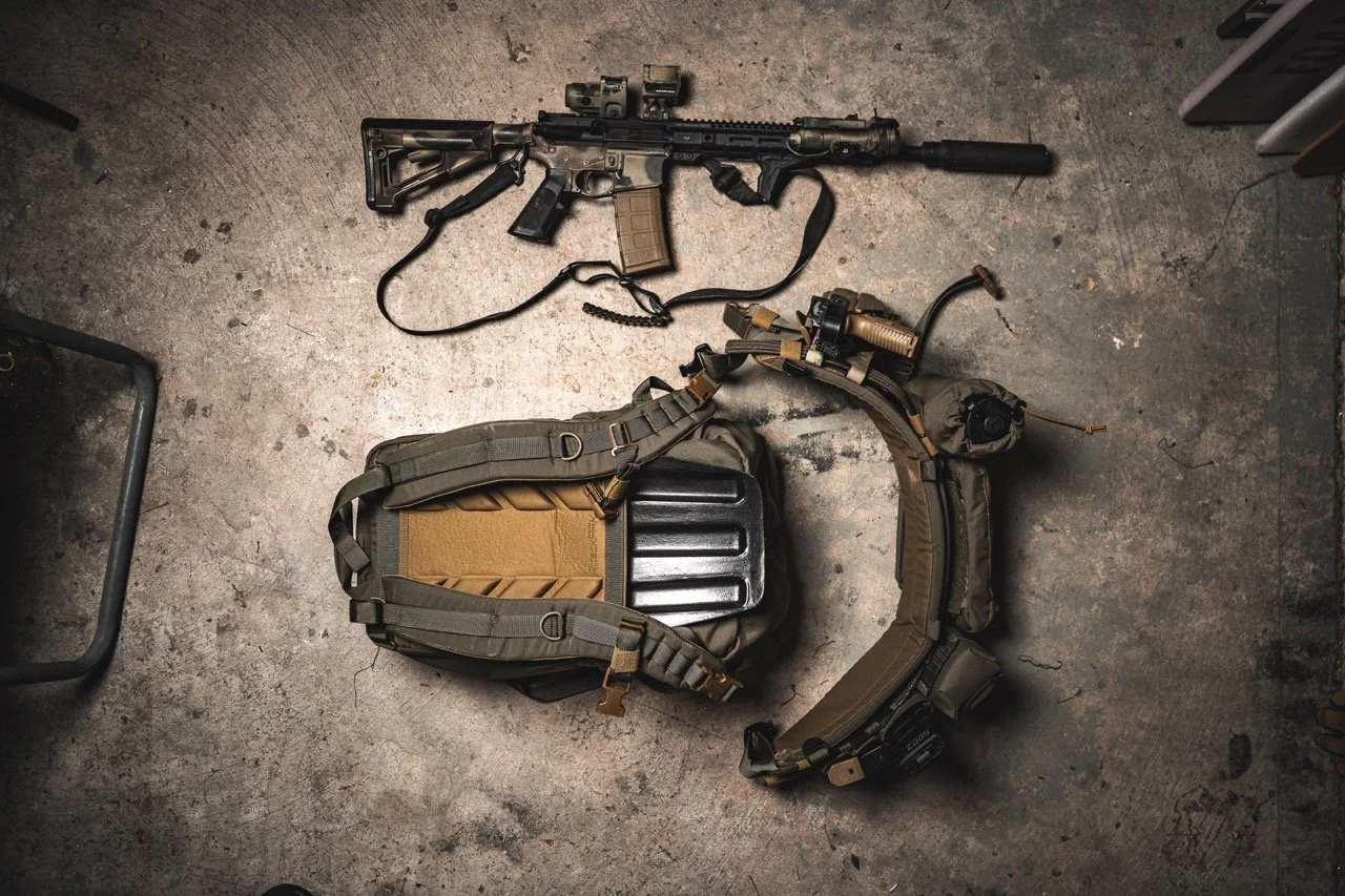 A tactical rifle with attachments and a tan backpack and belt laid out on a concrete floor.