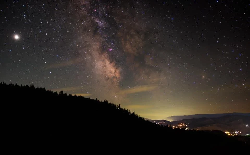 Night sky filled with stars and the Milky Way galaxy above a silhouetted landscape with distant lights.