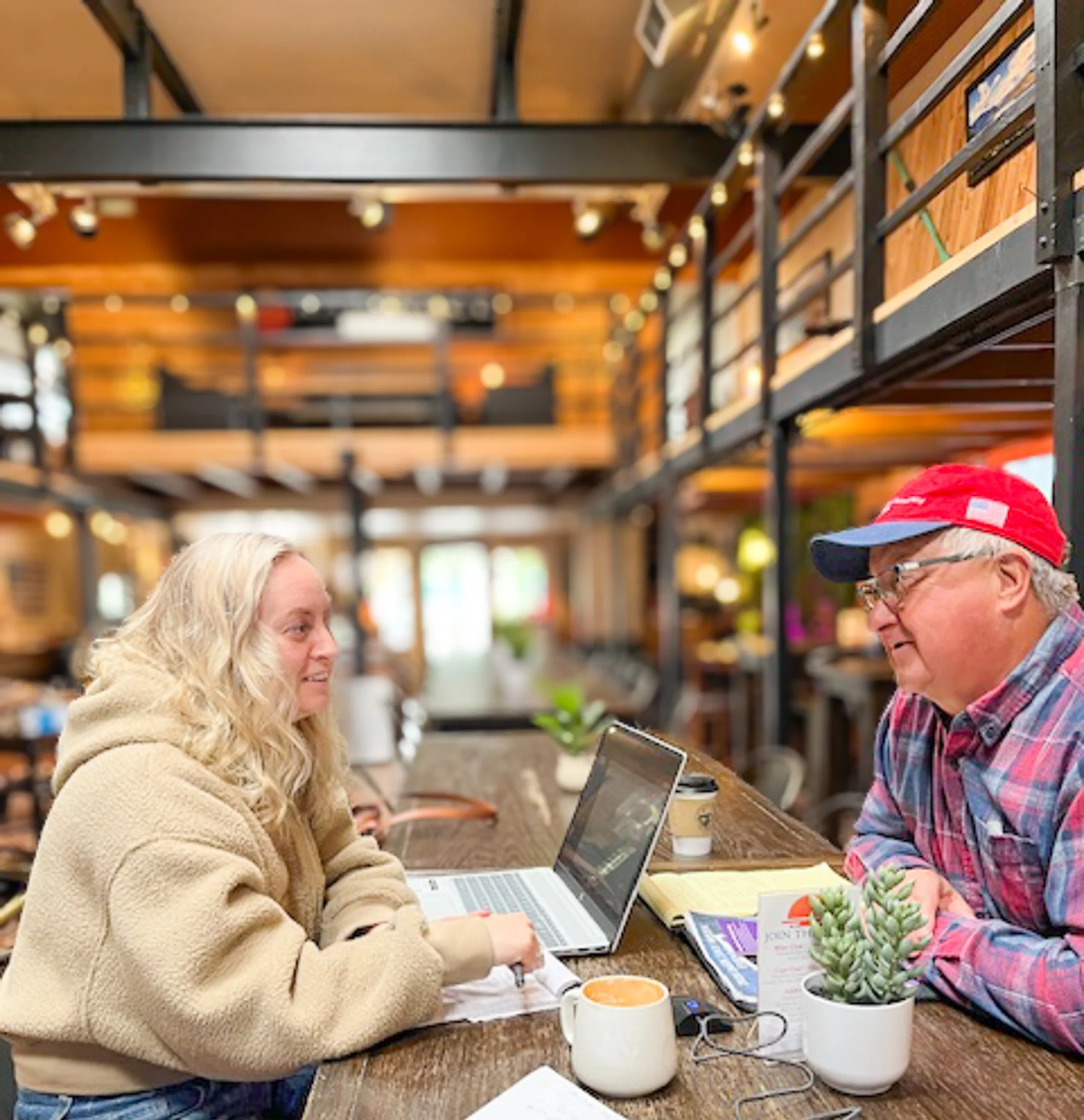 Two women sitting at a wooden table in a cozy indoor setting, engaged in conversation. They are surrounded by a laptop, a laptop, a book, a plant, and coffee cups. The background features warm lighting and wooden decor.