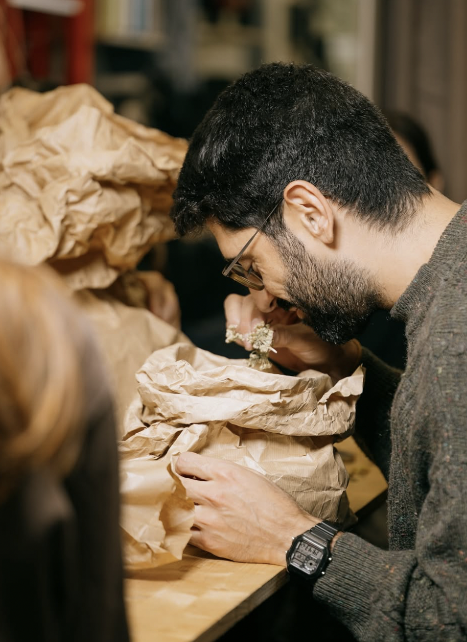 Un homme brun portant des lunettes et un pull gris examine une plante ou une fleur dans un sac en papier, entouré de plusieurs sacs en papier empilés.