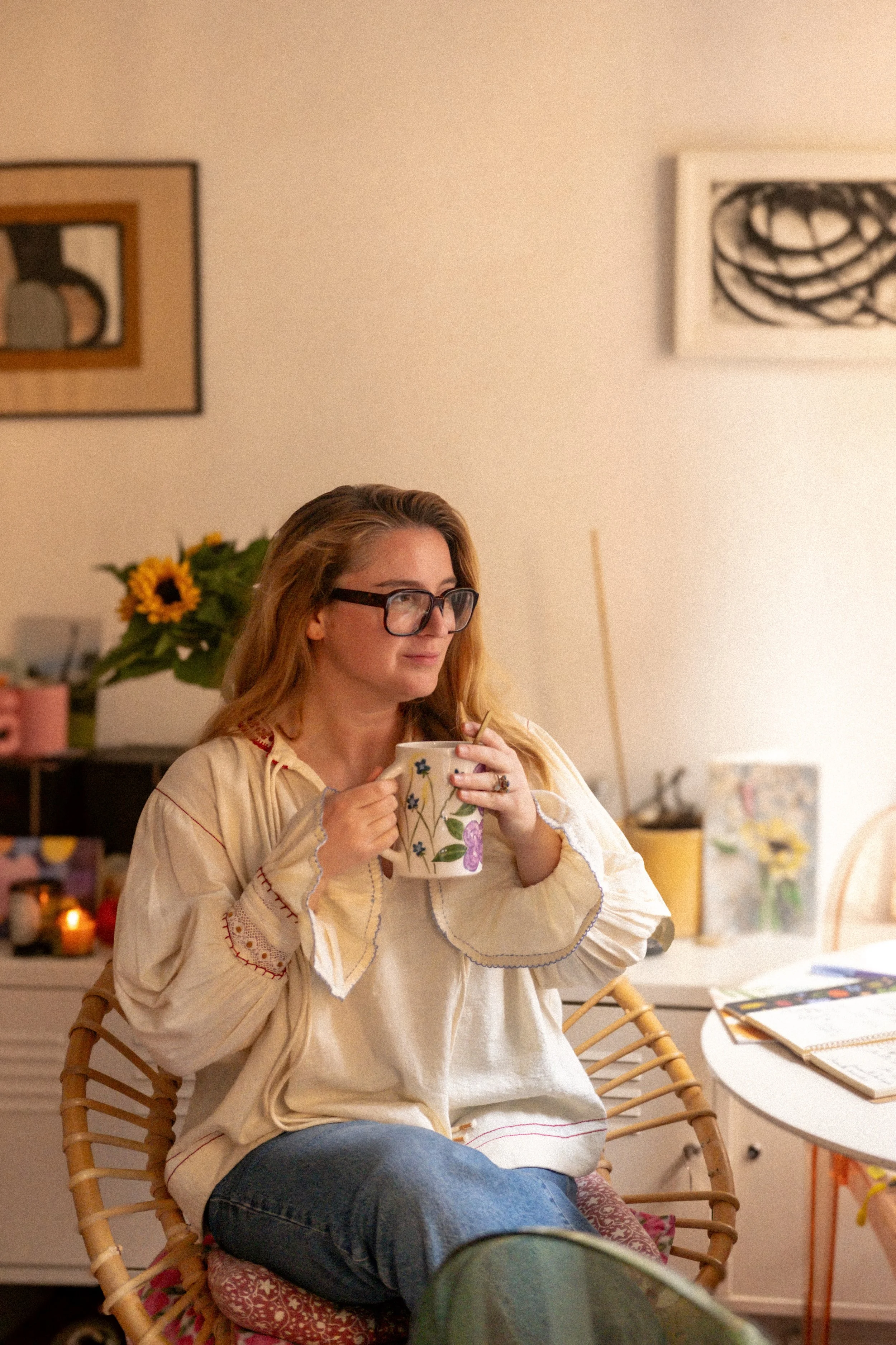 Une femme aux cheveux roux, portant des lunettes épaisses, sirote dans une tasse décorée de fleurs, assise sur une chaise en rotin dans un intérieur chaleureux, avec des tableaux modernes accroché au mur, une table à côté avec des magazines et une lumière douce.