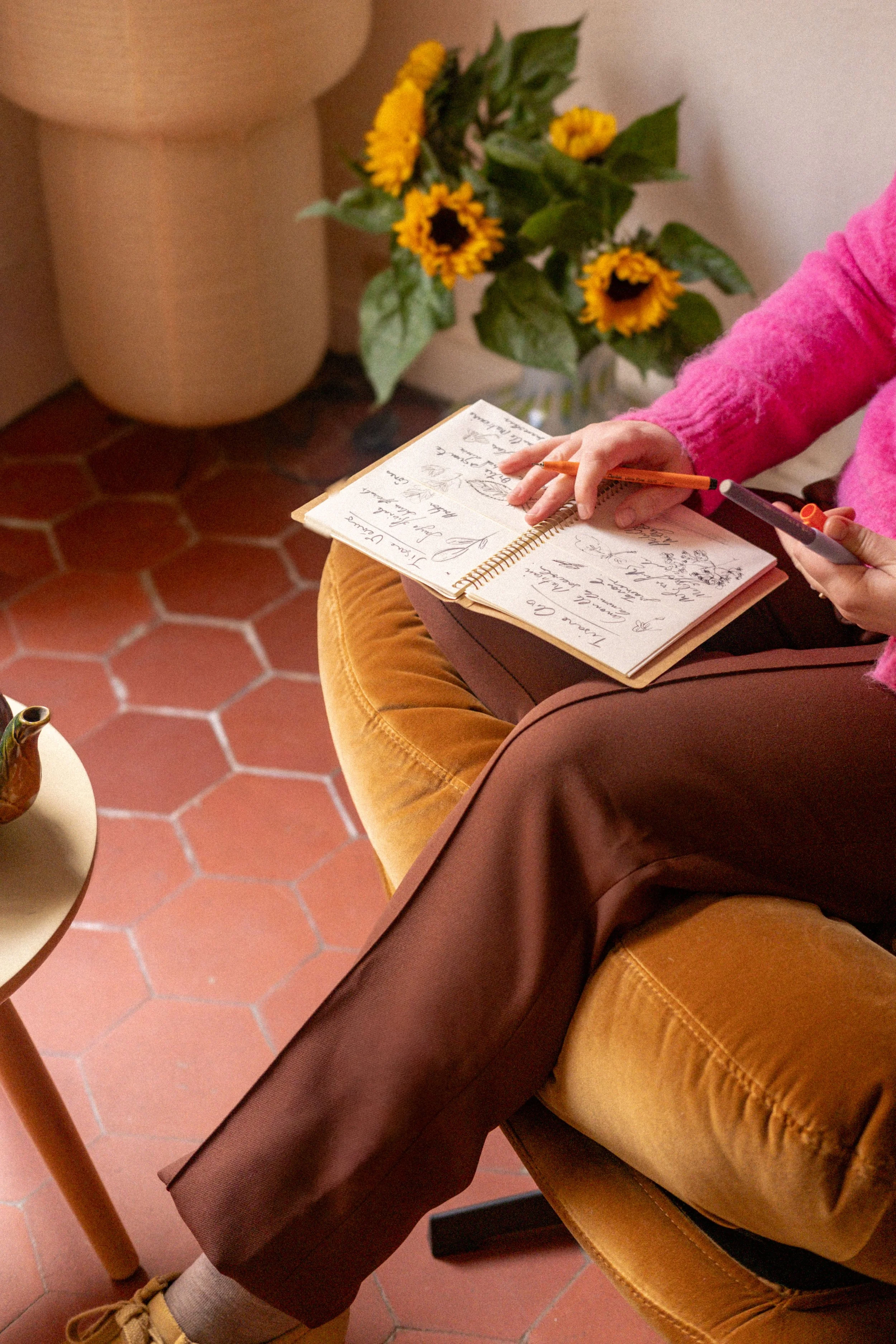 Une personne assise sur un fauteuil, en train d'écrire dans un carnet avec un stylo à la main, portant un pull rose et des pantalons marron, avec des fleurs de tournesol en arrière-plan sur une table.
