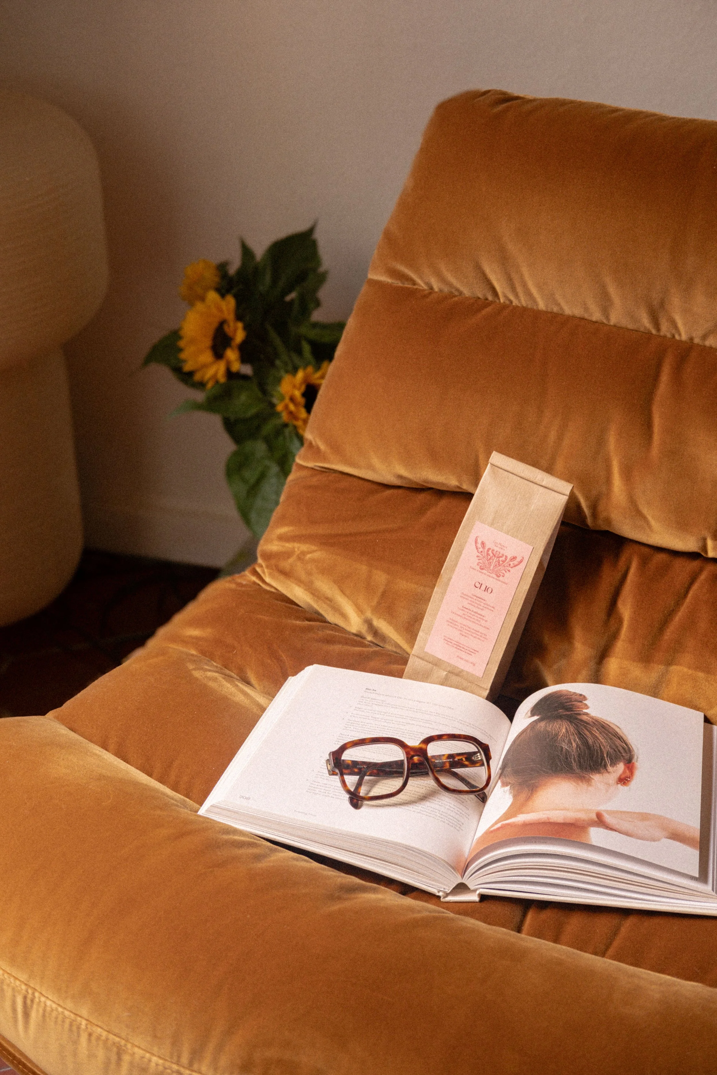 Une chaise orange en velours avec un livre ouvert, une paire de lunettes et un sachet de thé ou café dessus, derrière un bouquet de tournesols.