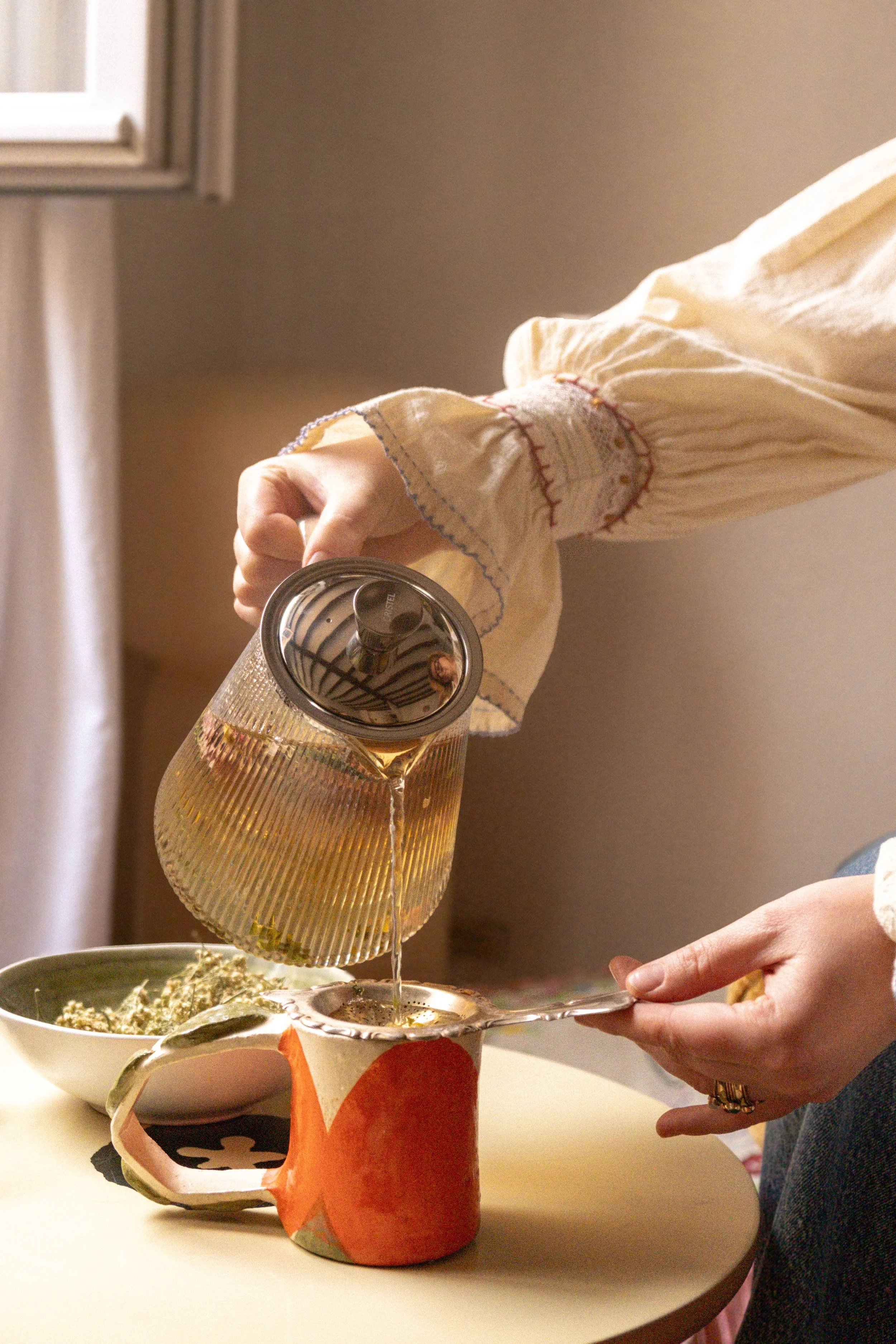 Une personne verse du thé dans une tasse en céramique orange avec un motif animalier, une assiette avec des plantes en arrière-plan, dans une pièce lumineuse.