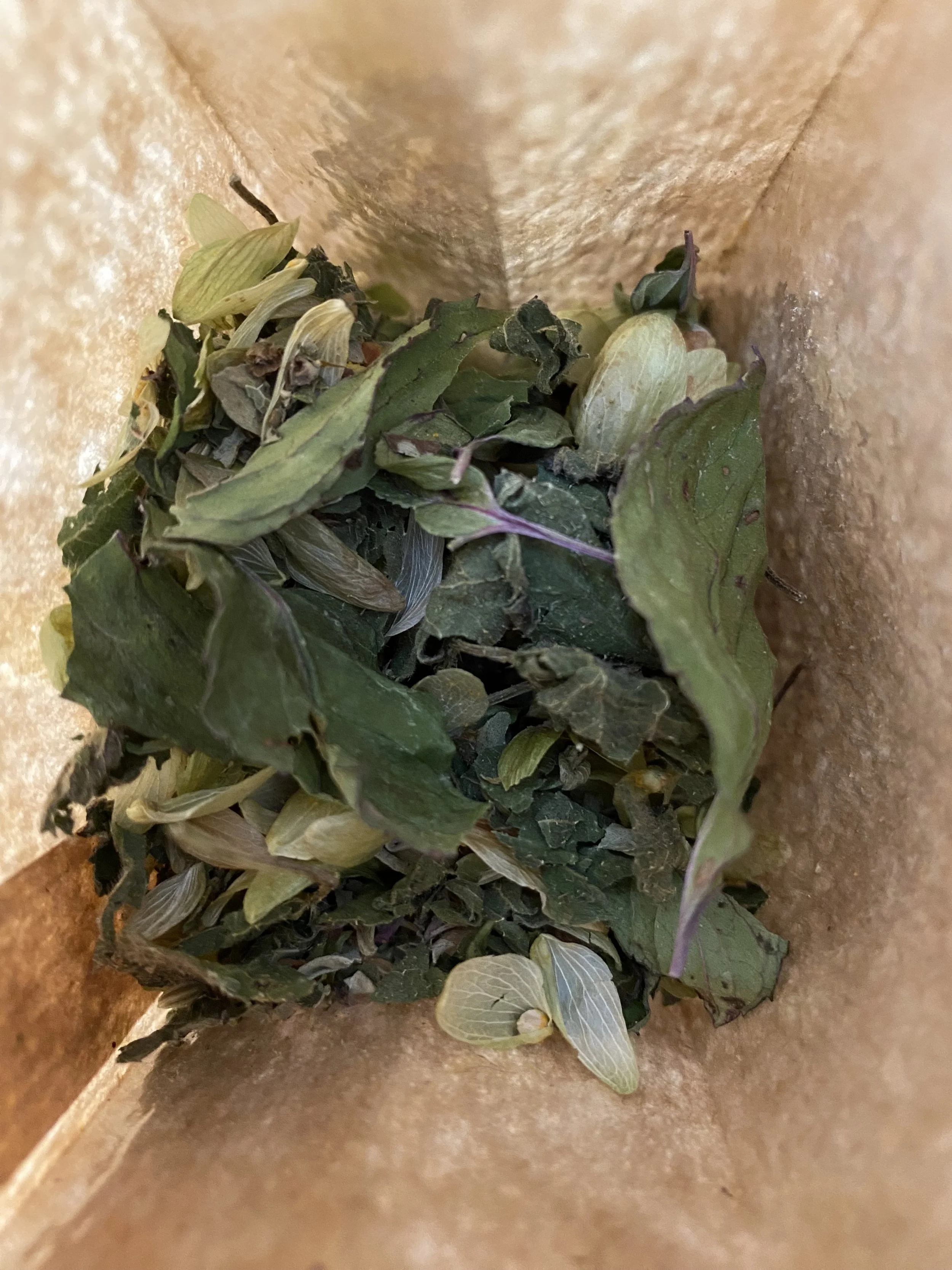 Une table avec des sachets de tisane, un carnet de notes jaune intitulé « Fredonner Vie », une bougie allumée, un livre intitulé « The Play » en anglais, et un plat contenant des herbes séchées.