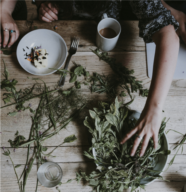 Une personne arrange des plantes et des feuilles sur une table en bois, avec une tasse de café, une assiette avec une portion de dessert, une fourchette, et un verre.