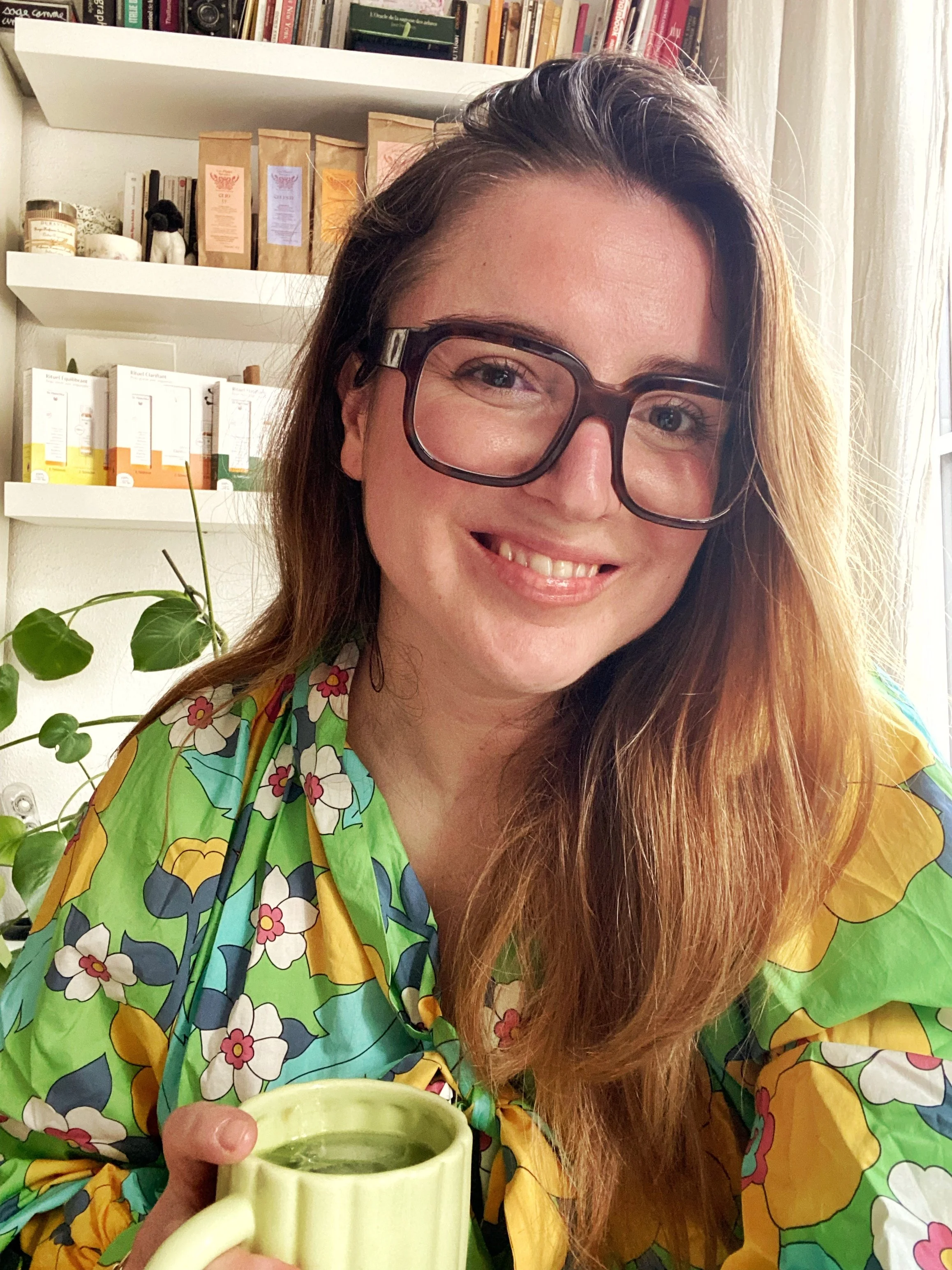Une femme souriante avec de longues cheveux roux, portant de grandes lunettes, vêtue d'une chemise à fleurs verte, jaune, et bleue, tenant une tasse de café ou de thé, dans un intérieur avec étagères et une plante verte en arrière-plan.