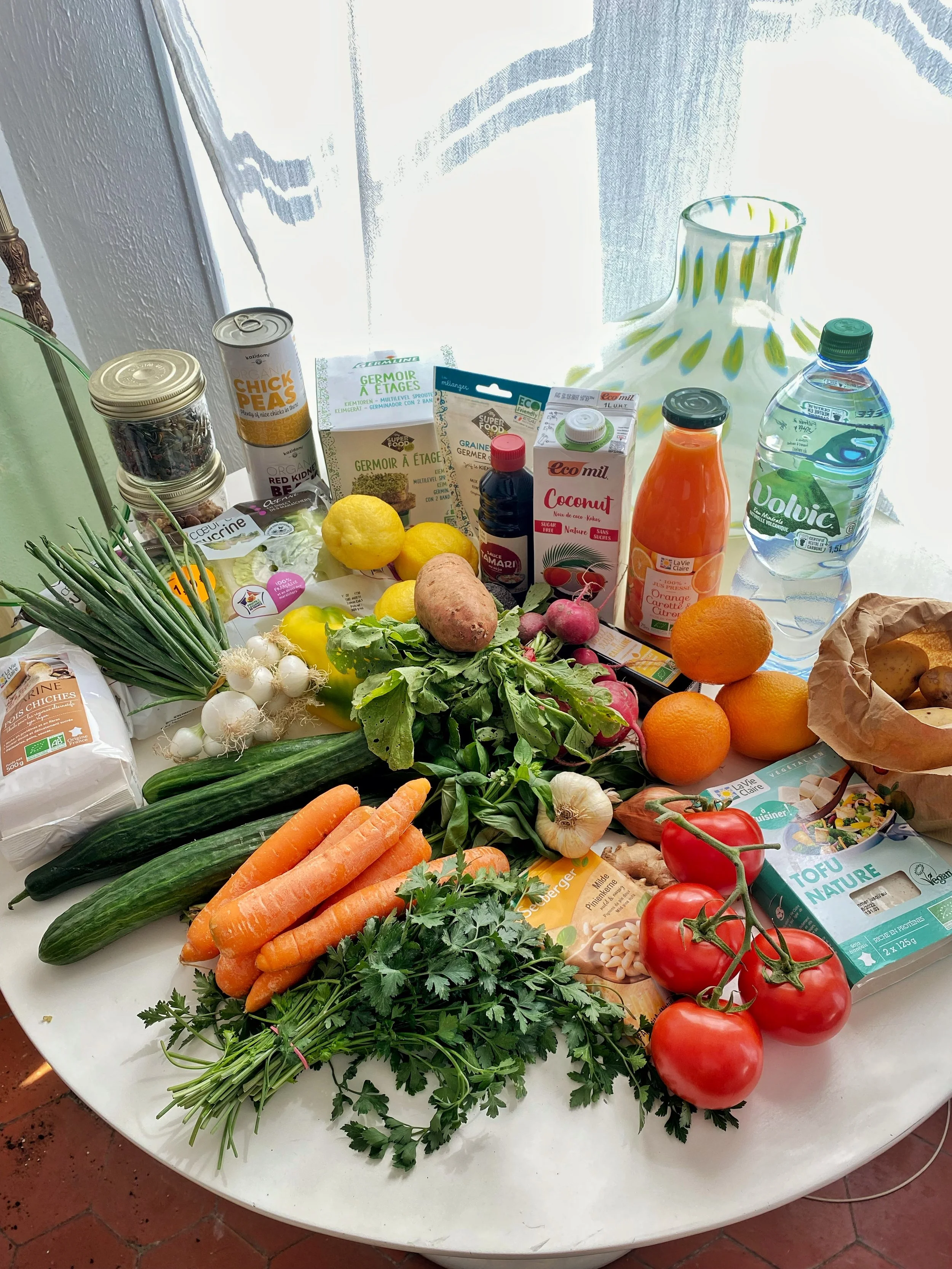 Une table chargée de divers produits alimentaires et légumes tels que des tomates, des carottes, des radis, des concombres, de l'ail, des citrons, des oranges, des pommes de terre, des oignons verts, et des aliments emballés, avec une cruche en verre et une bouteille d'eau en arrière-plan.