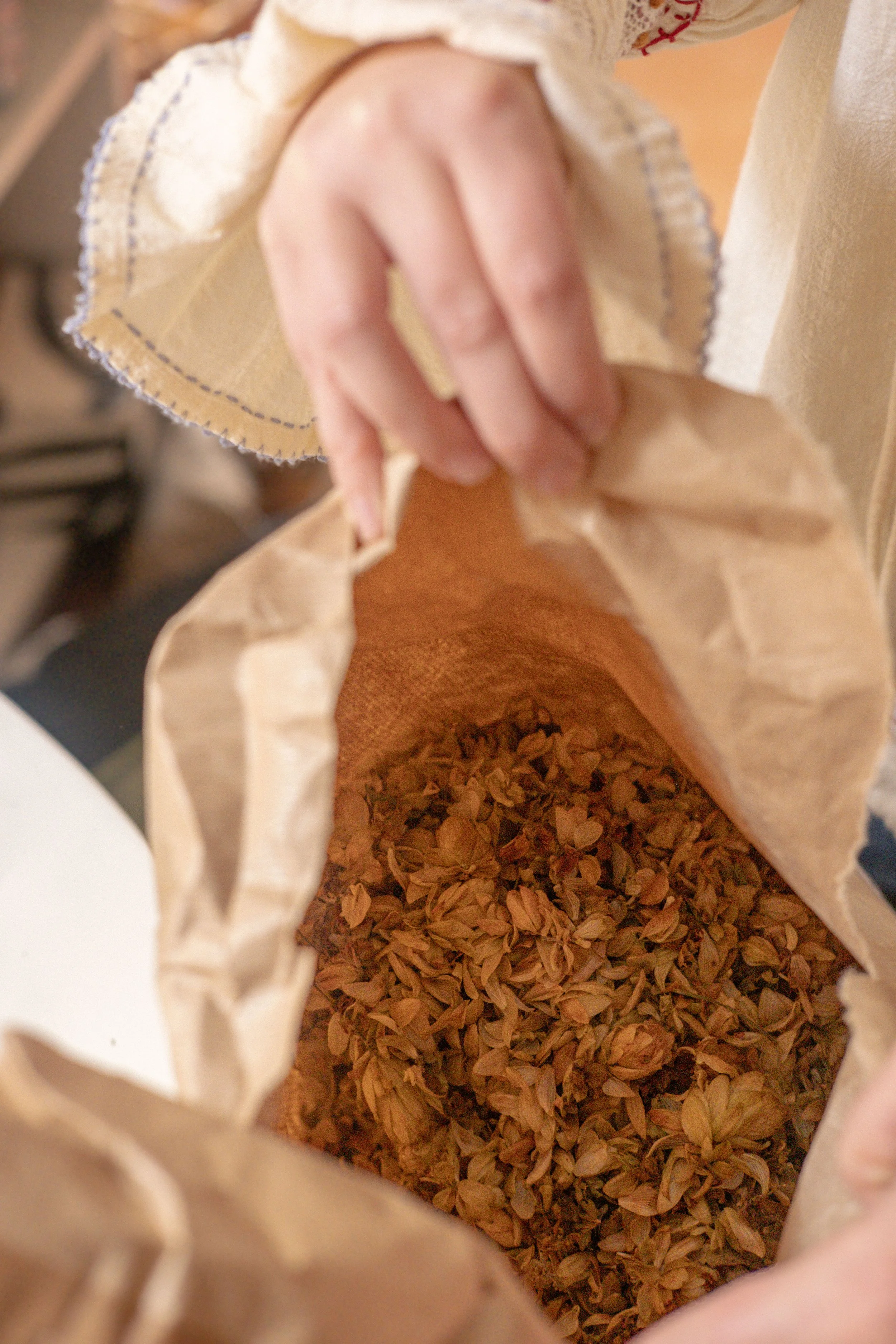 Main tenant un sac en papier rempli de fleurs séchées ou d'herbes, avec une main qui tient le sac en haut et une autre qui ouvre le sac.
